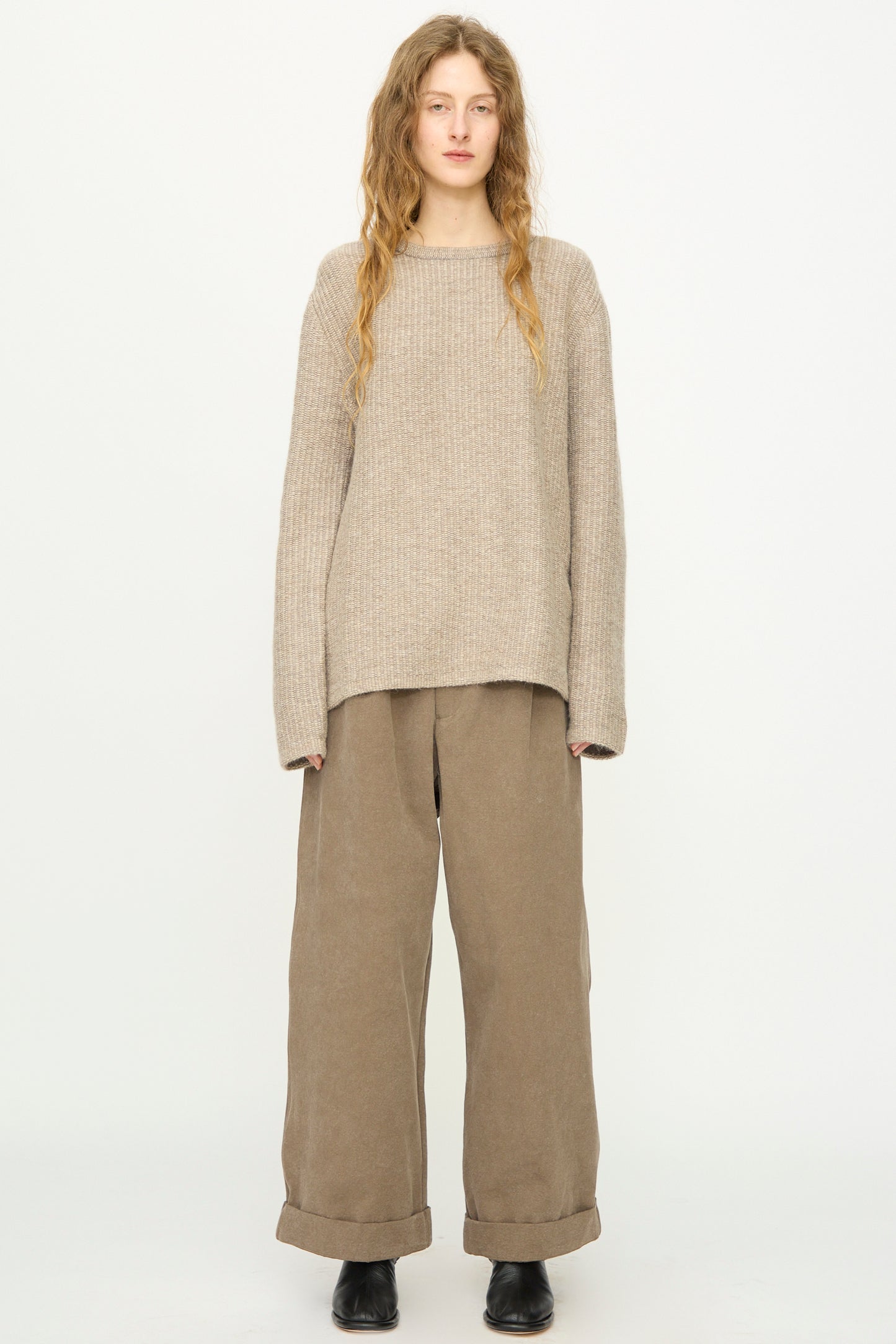 A person stands against a plain background wearing Lauren Manoogian's Knitweave Pullover in Oatmeal, paired with wide-legged brown pants and black shoes.