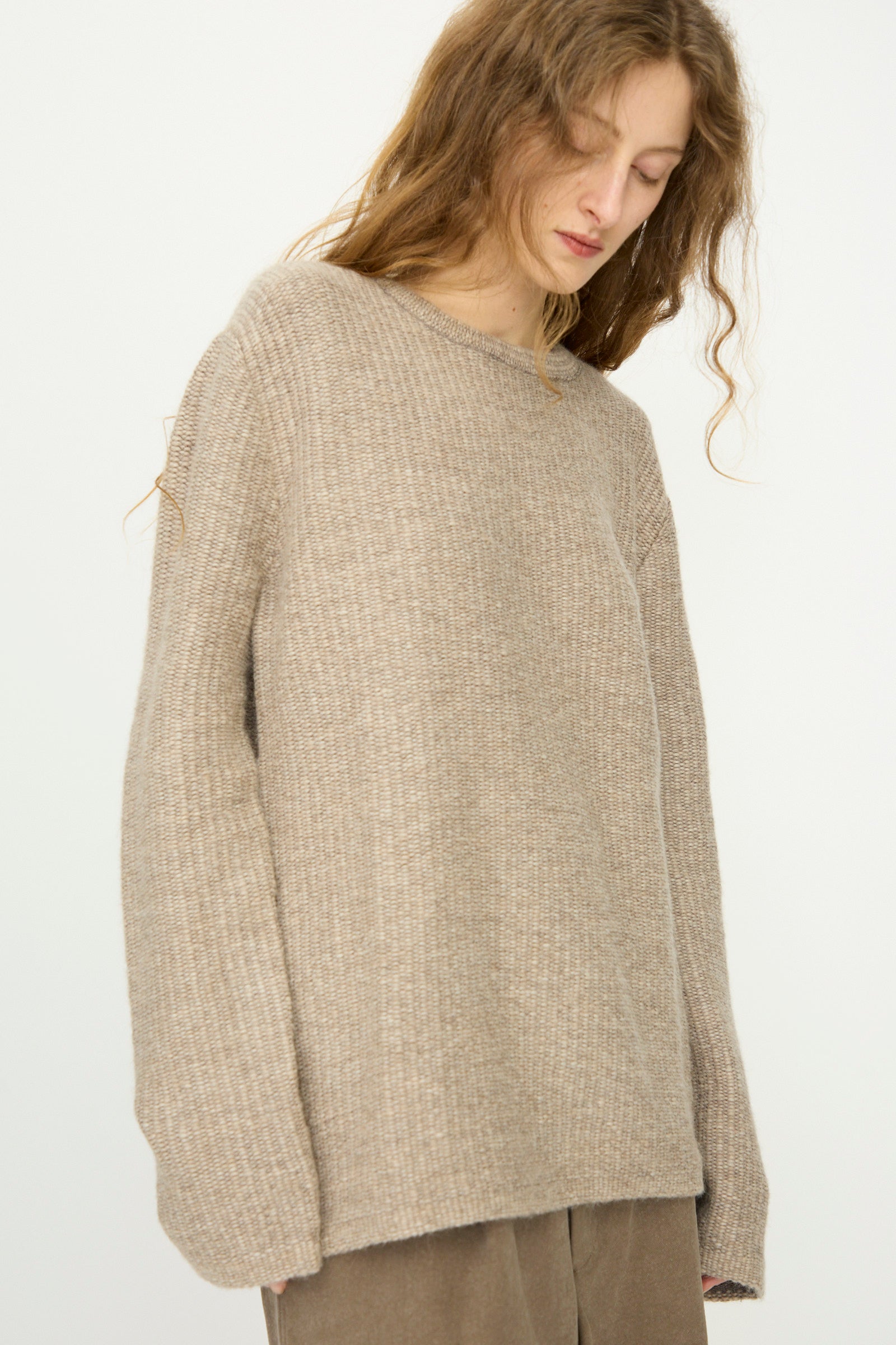 Model wears the Lauren Manoogian Knitweave Pullover in Oatmeal—a loose-fitting, beige baby alpaca sweater—with brown pants, standing against a plain light background and looking downward.