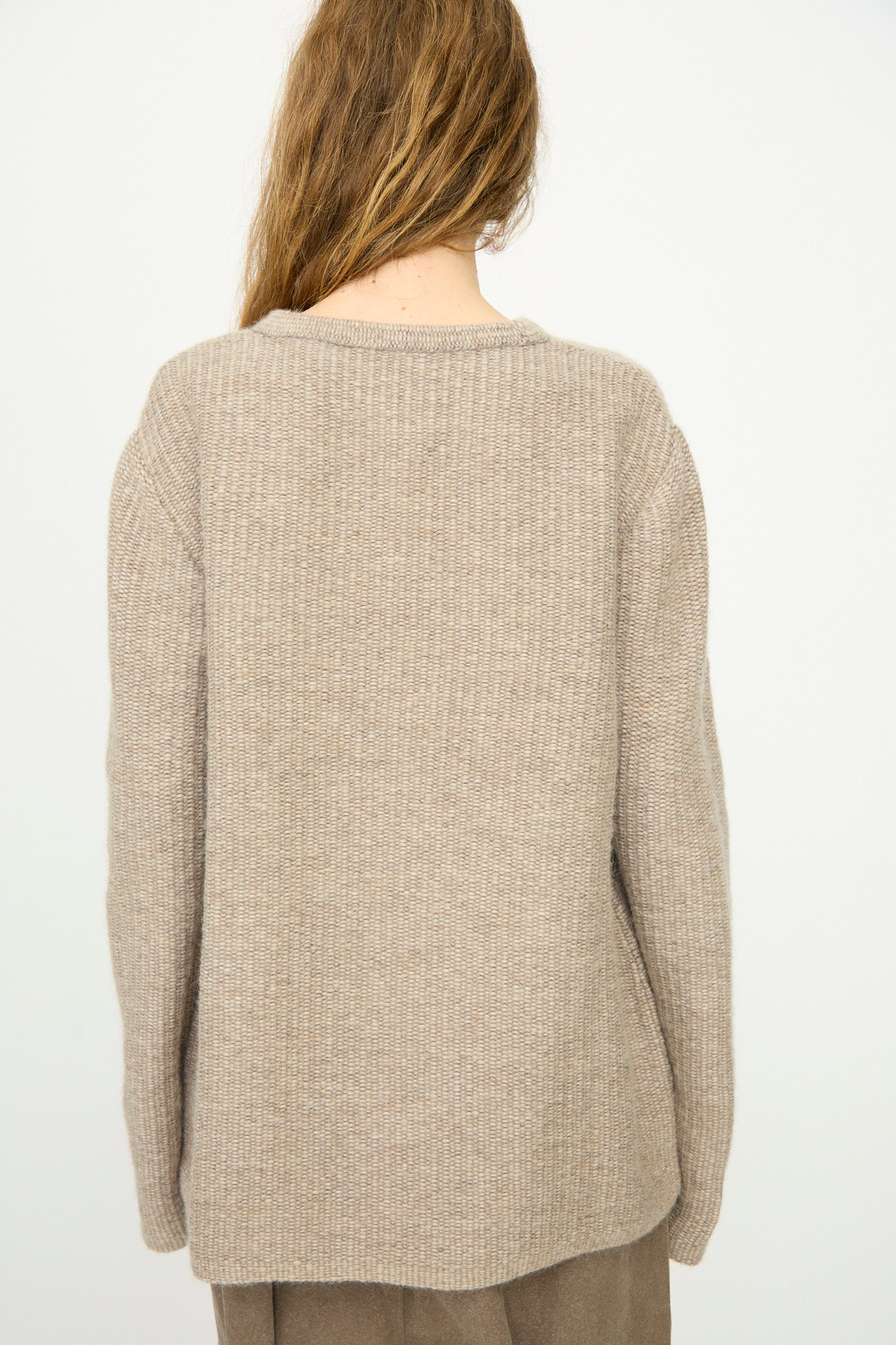 A person with long, light brown hair is seen from behind wearing the Lauren Manoogian Knitweave Pullover in Oatmeal and brown pants against a plain light background.