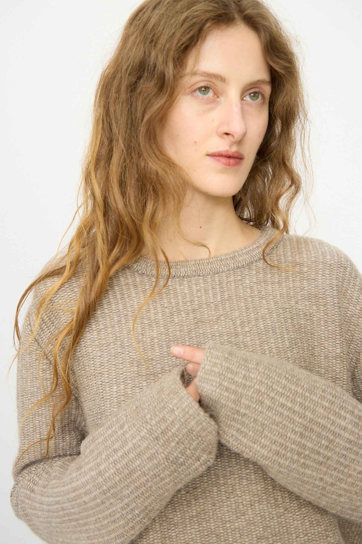 A woman with long wavy hair wears the Lauren Manoogian Knitweave Pullover in Oatmeal, standing against a plain light background and gazing slightly to the side.