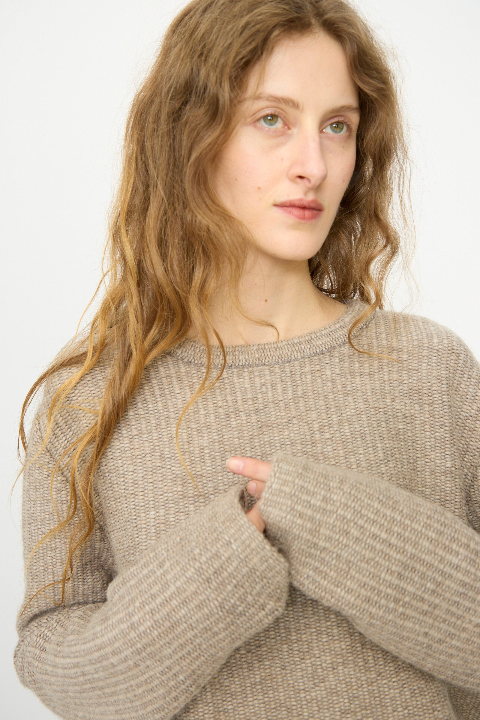 A woman with long wavy hair wears the Lauren Manoogian Knitweave Pullover in Oatmeal, standing against a plain light background and gazing slightly to the side.