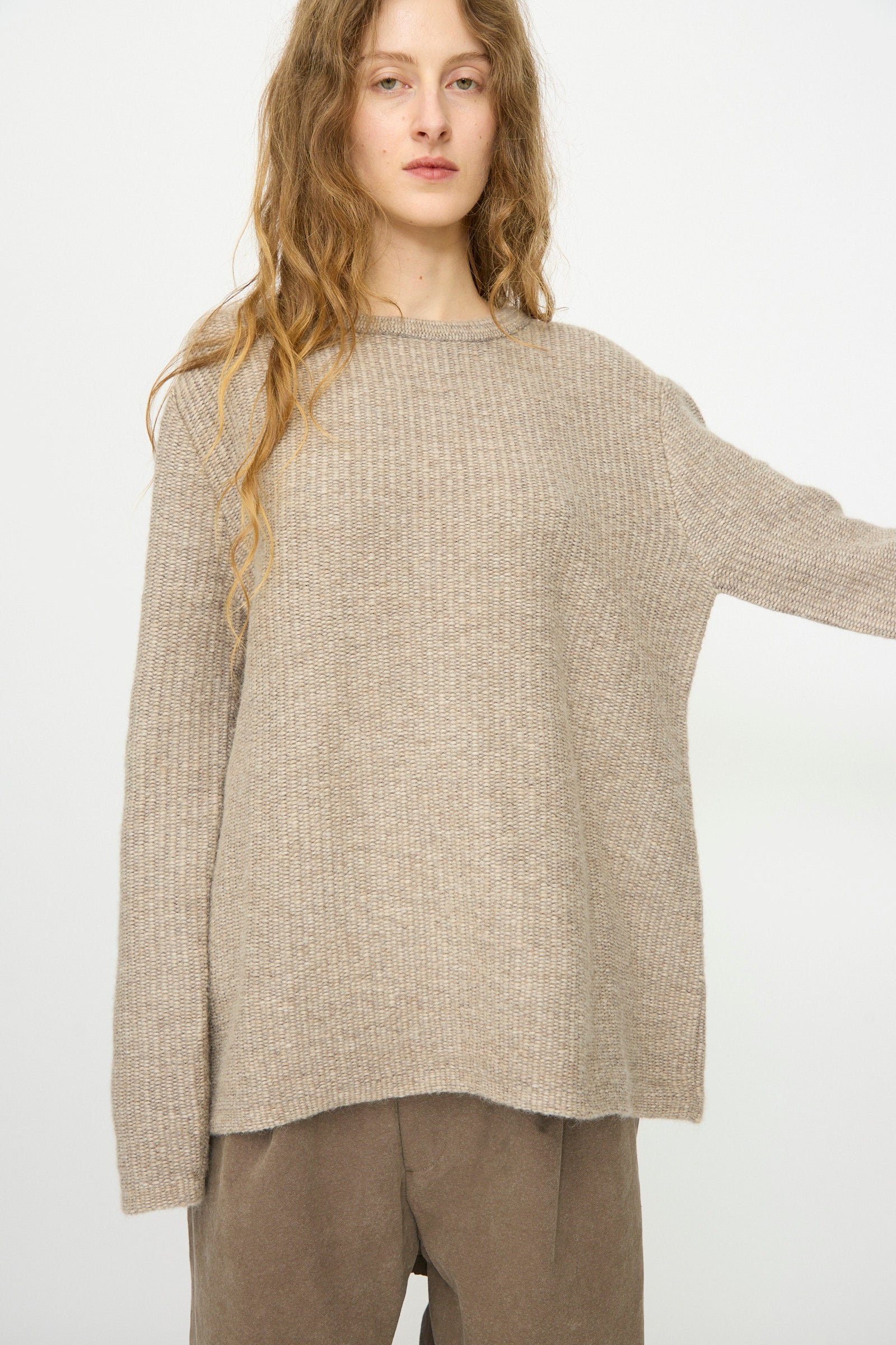 A person with long wavy hair wears the Lauren Manoogian Knitweave Pullover in Oatmeal and brown pants, standing against a plain white background.