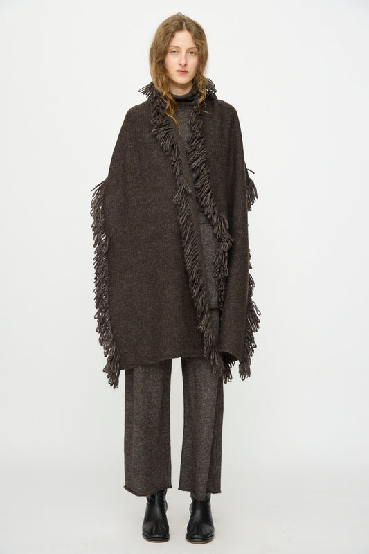 A person faces forward wearing the Lauren Manoogian Knitweave Wrap in Blackened over matching wide-leg pants and black ankle boots, accessorized with a hand-loomed scarf, against a plain white background.