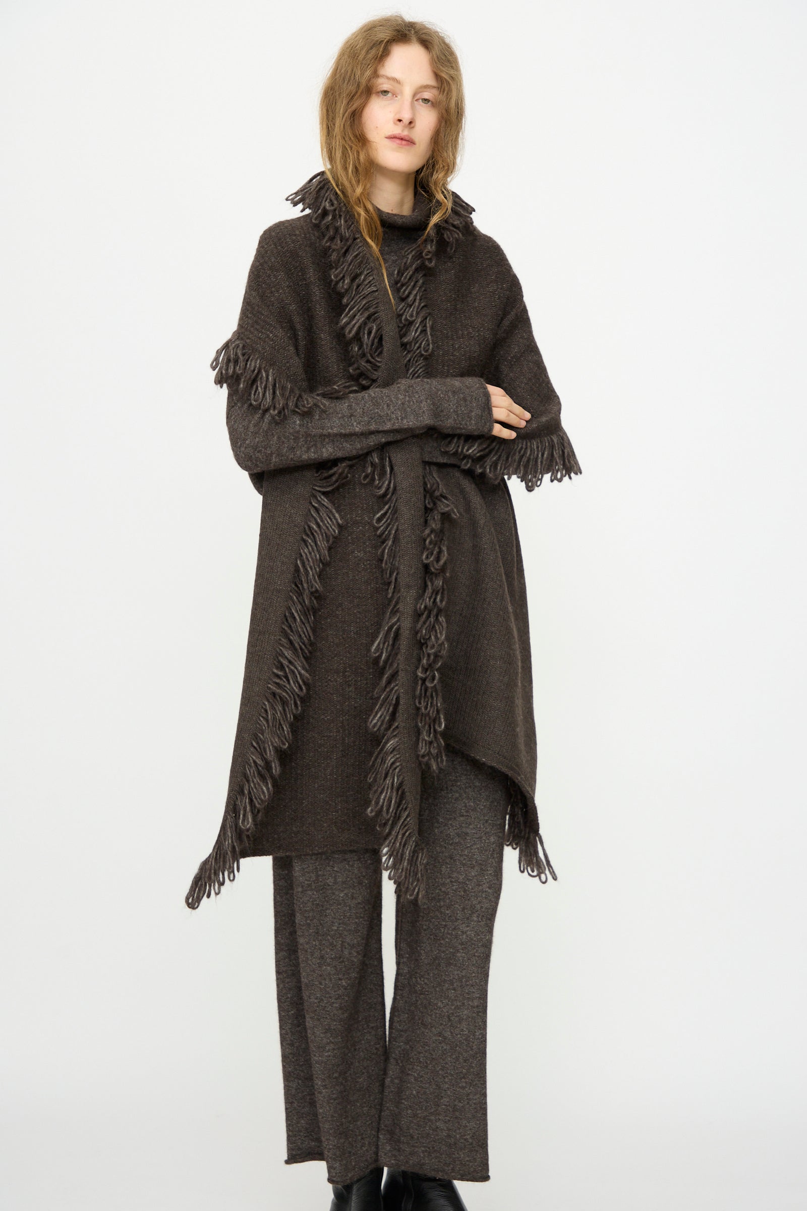 A person stands against a white background wearing Lauren Manoogian’s Knitweave Wrap in Blackened, styled over matching knit pants and a black turtleneck, with a hand-loomed scarf.