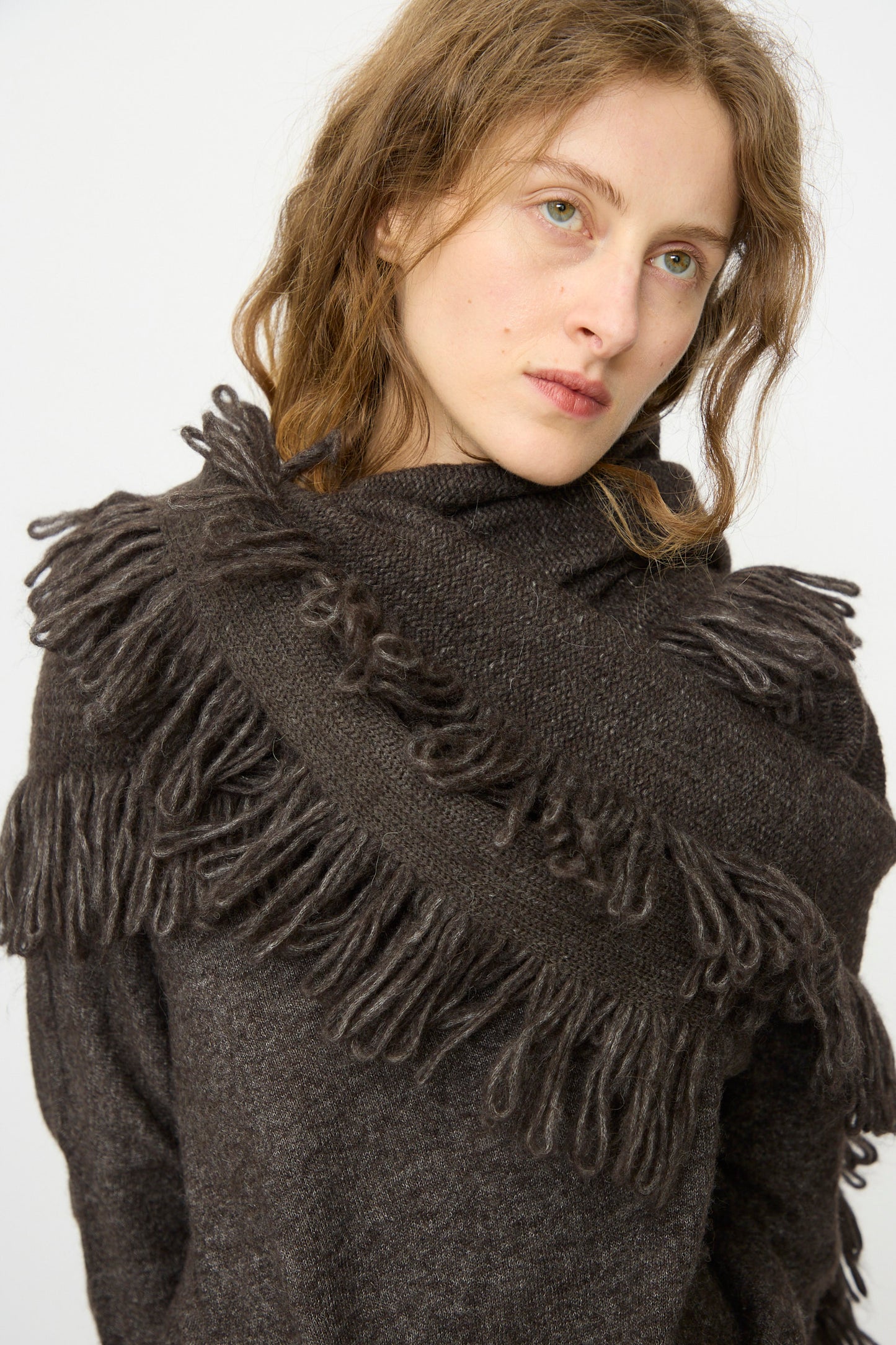 A person with wavy light brown hair wears the Lauren Manoogian Knitweave Wrap in Blackened and a dark brown knit sweater with fringe, standing against a plain white background.