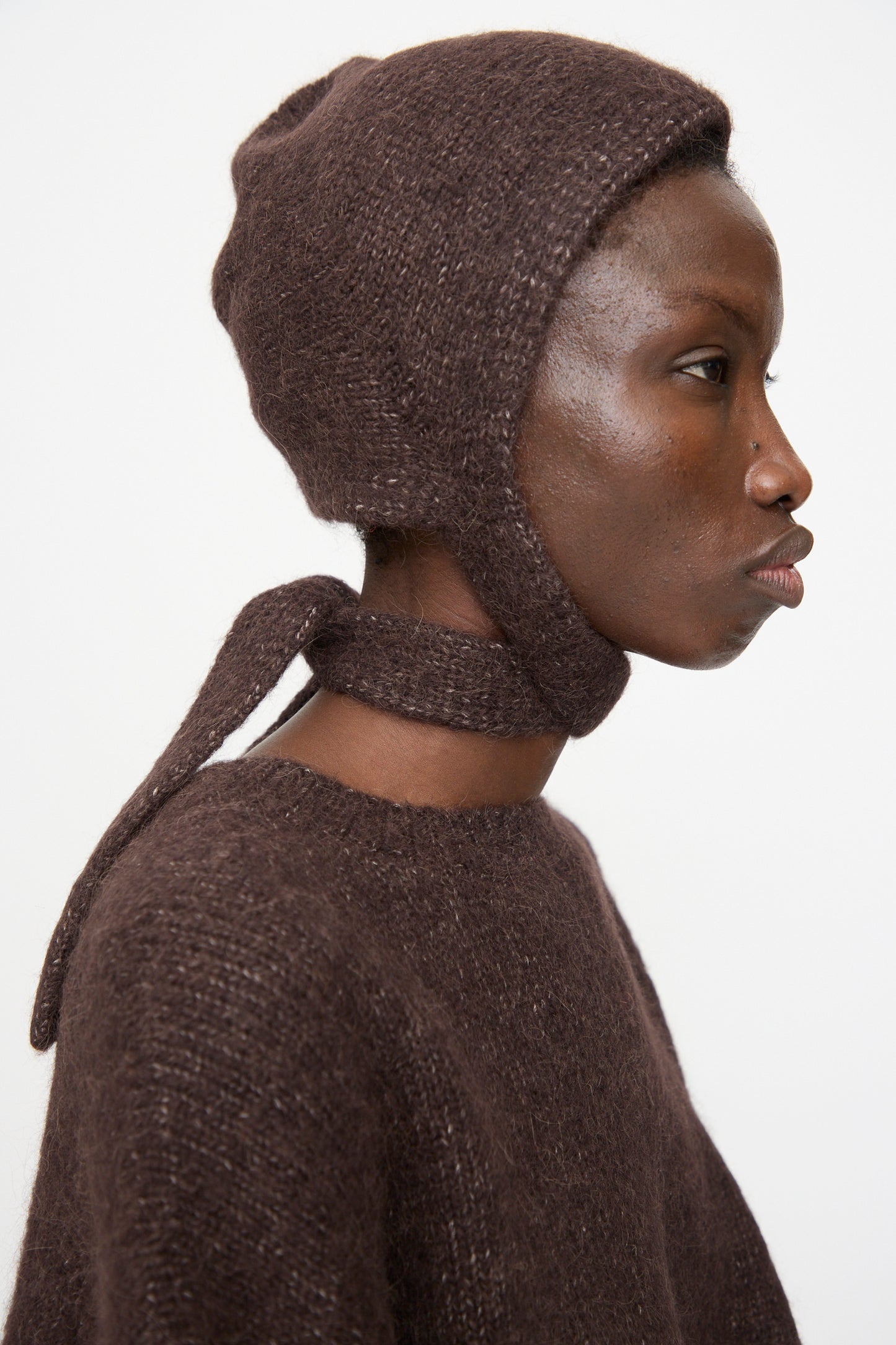 A person in profile wears the Lauren Manoogian Loft Bonnet in Soil, a fitted suri alpaca blend with ties, paired with a matching brown knit top, all made in Peru, set against a plain white background.