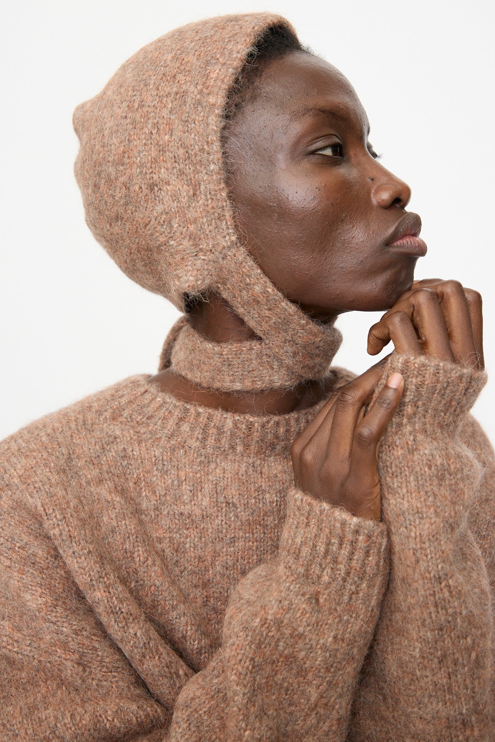A person in a Lauren Manoogian Loft Bonnet in Spice and a textured brown suri alpaca blend sweater, made in Peru, gazes to the side with hand on chin against a white background.