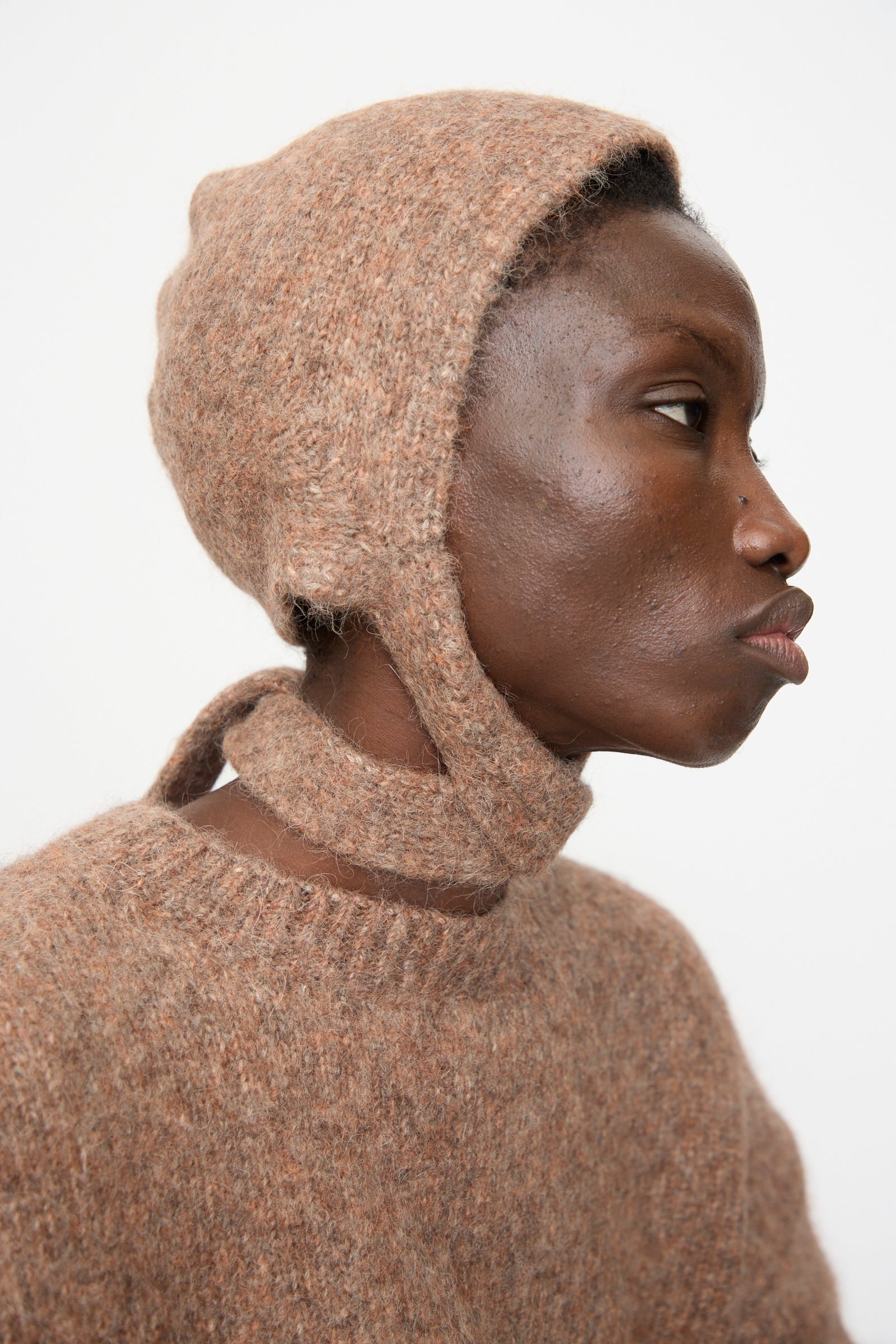 A person in profile wears the Lauren Manoogian Loft Bonnet in Spice, a fitted suri alpaca blend bonnet with chin strap, paired with a matching sweater. Both pieces are made in Peru and set against a plain white background.