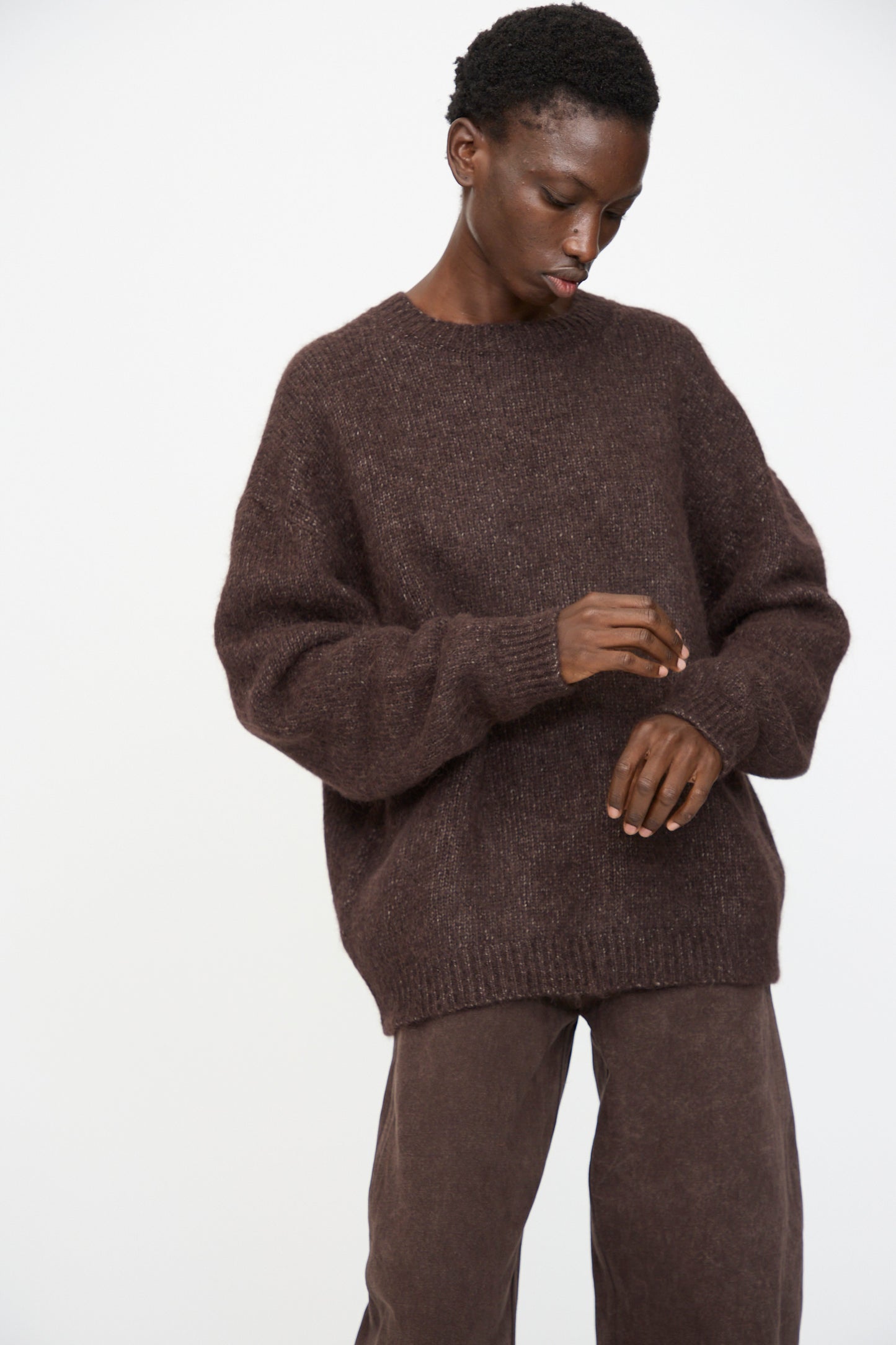 A person wears the Lauren Manoogian Loft Crewneck in Soil with matching brown pants, standing against a plain light background and looking down with hands slightly raised.
