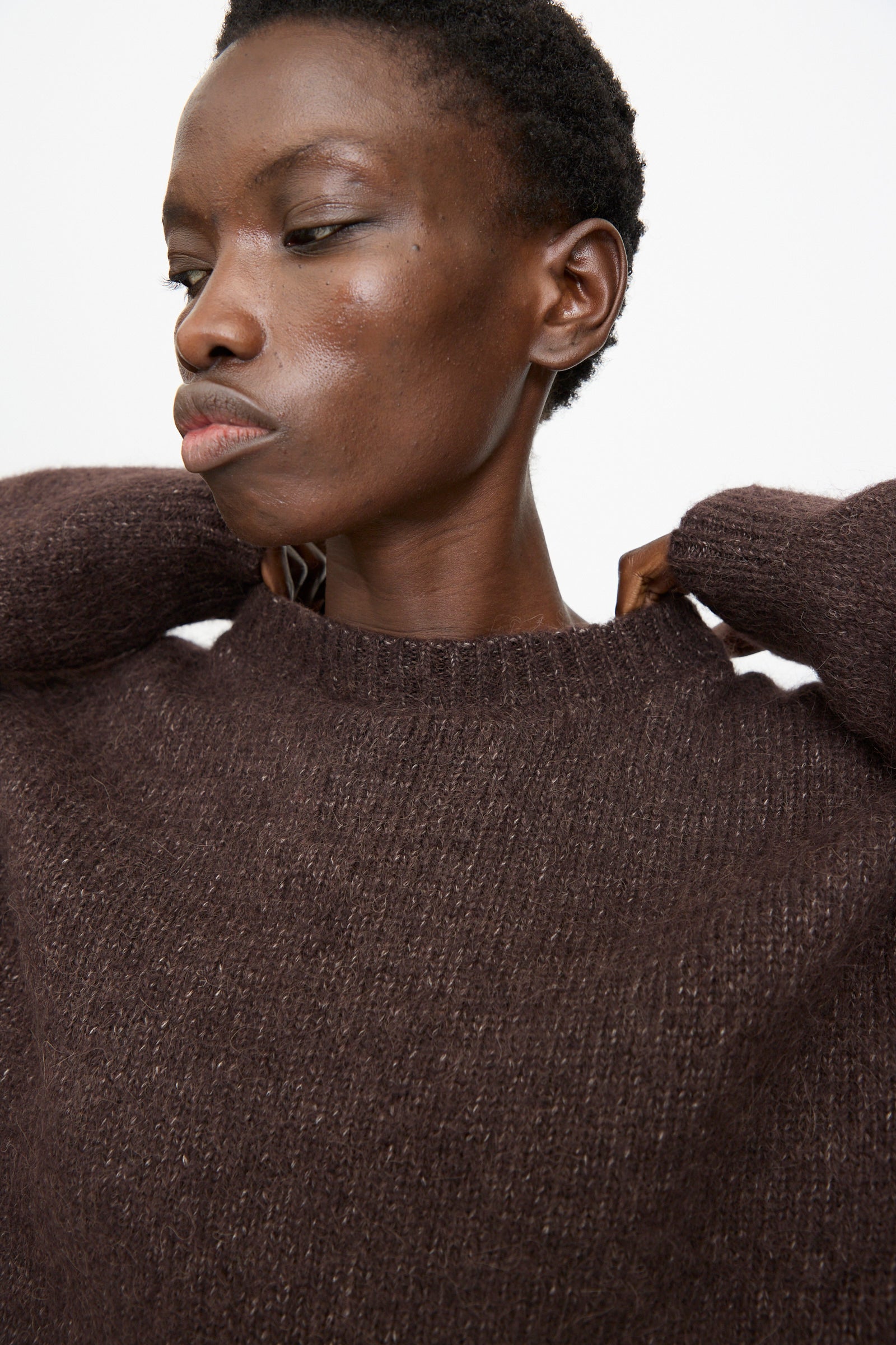 A person wearing the Lauren Manoogian Loft Crewneck in Soil, looking to the side with hands near the collar against a plain light background.