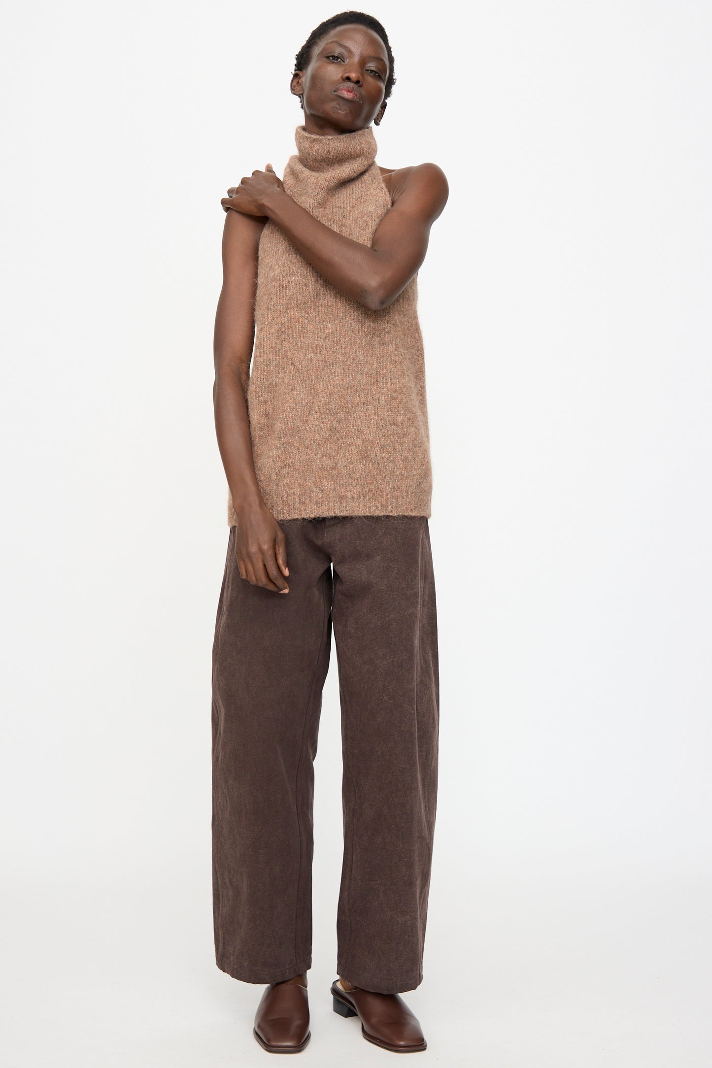 A person stands against a white background wearing the Lauren Manoogian Loft Halter in Spice, paired with wide-leg brown pants and brown shoes, with one arm resting on their shoulder.