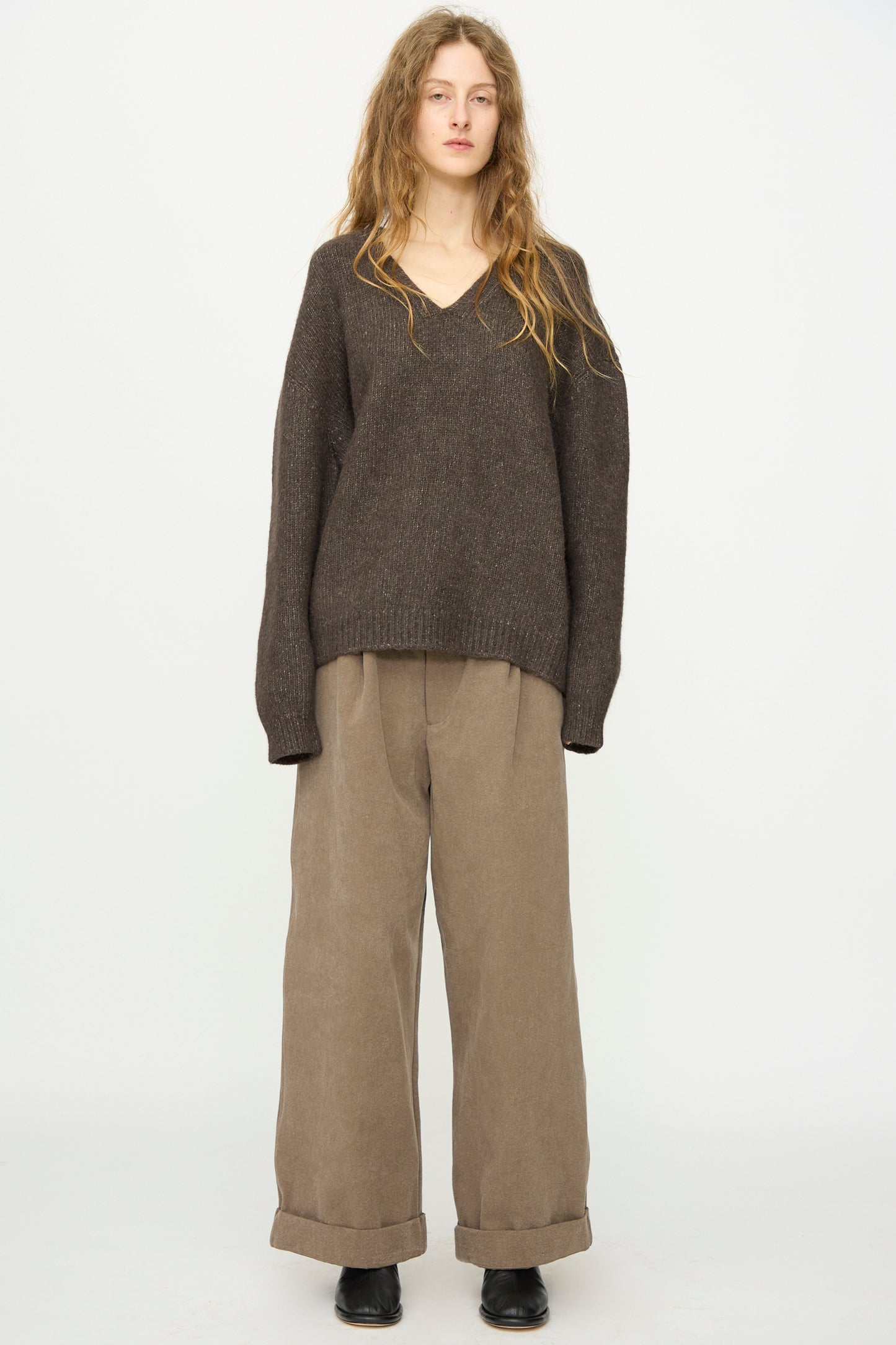 A person stands against a plain background wearing the Lauren Manoogian Loft V Neck in Coal, relaxed fit, paired with wide-legged beige cuffed pants and black shoes.