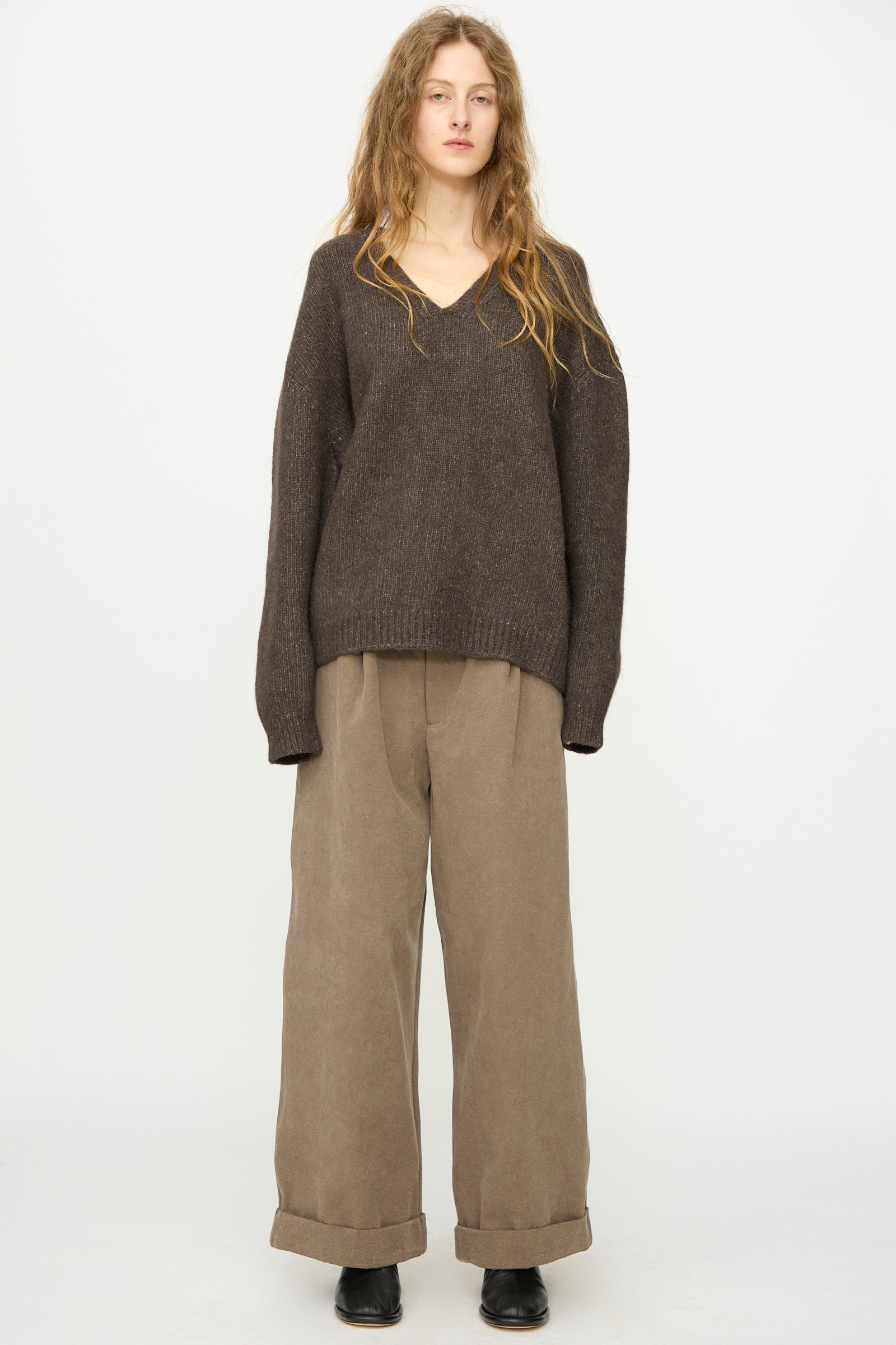 A person stands against a plain background wearing the Lauren Manoogian Loft V Neck in Coal, relaxed fit, paired with wide-legged beige cuffed pants and black shoes.