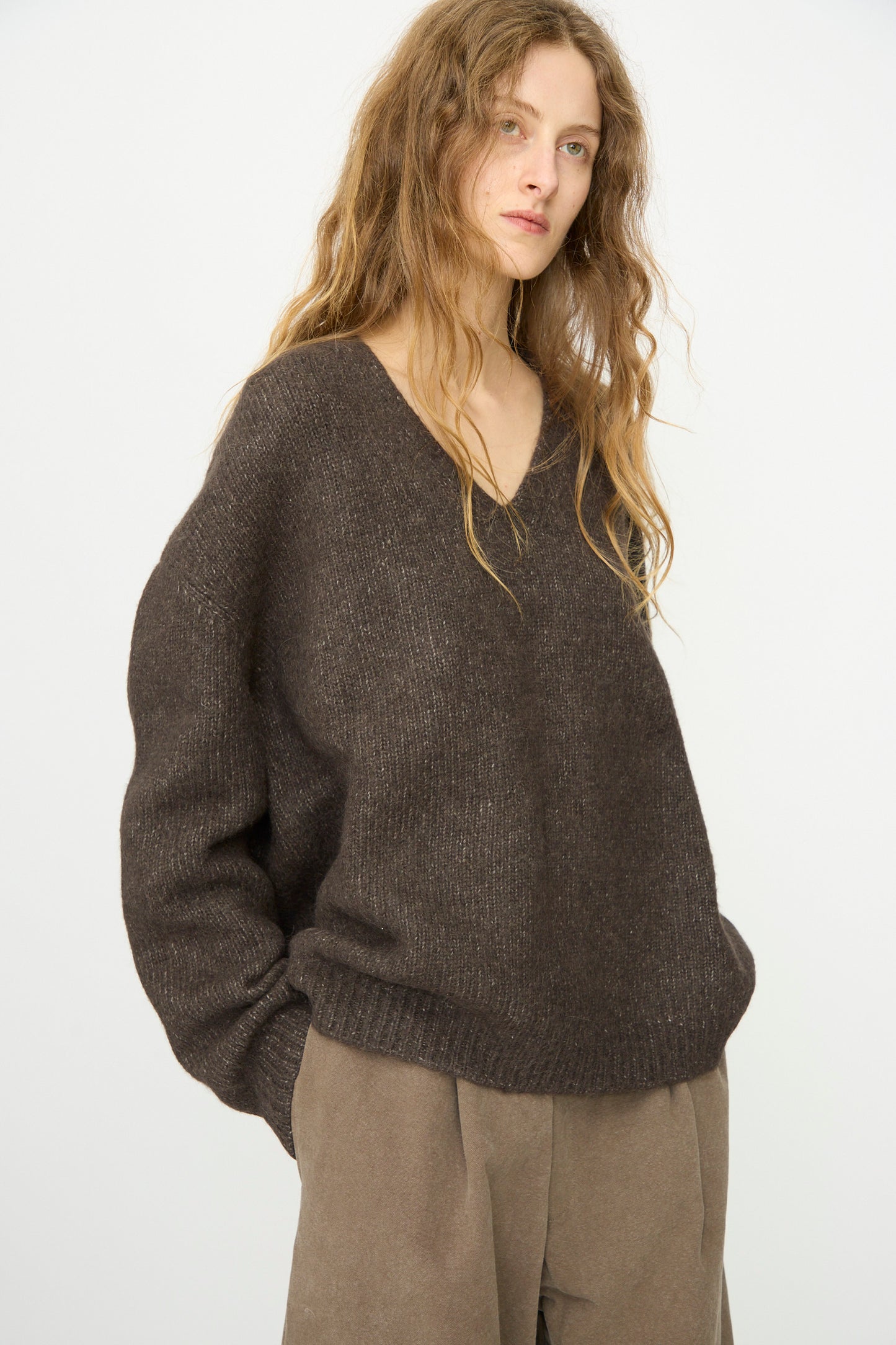 A person with long wavy hair wears the Lauren Manoogian Loft V Neck in Coal and taupe pants, standing against a plain white background.