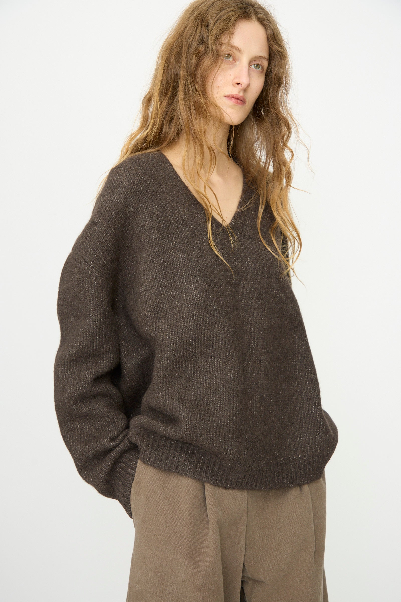 A person with long wavy hair wears the Lauren Manoogian Loft V Neck in Coal and taupe pants, standing against a plain white background.