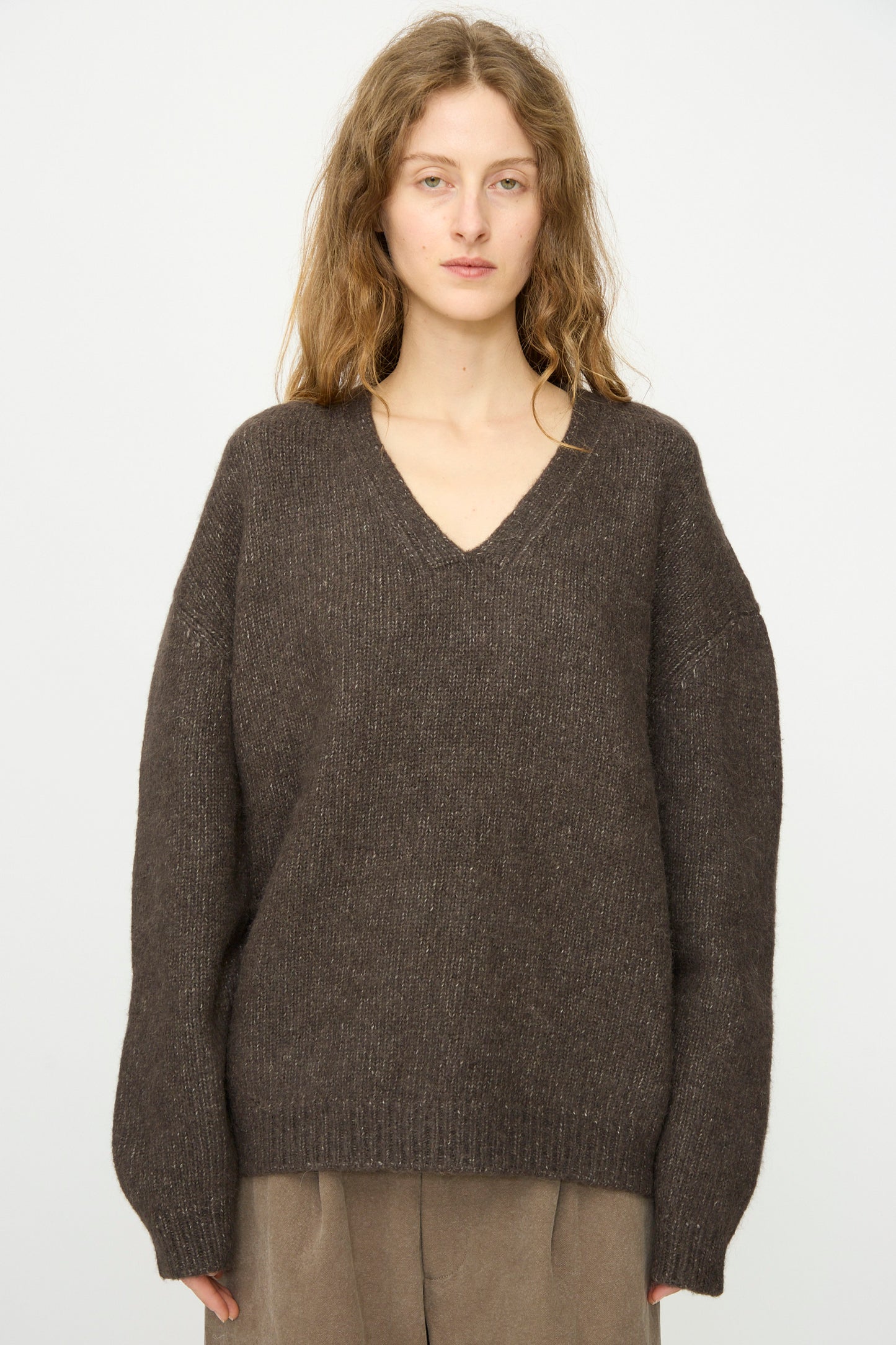 Woman with long, wavy hair wears the Lauren Manoogian Loft V Neck in Coal and beige pants, standing against a plain light background.