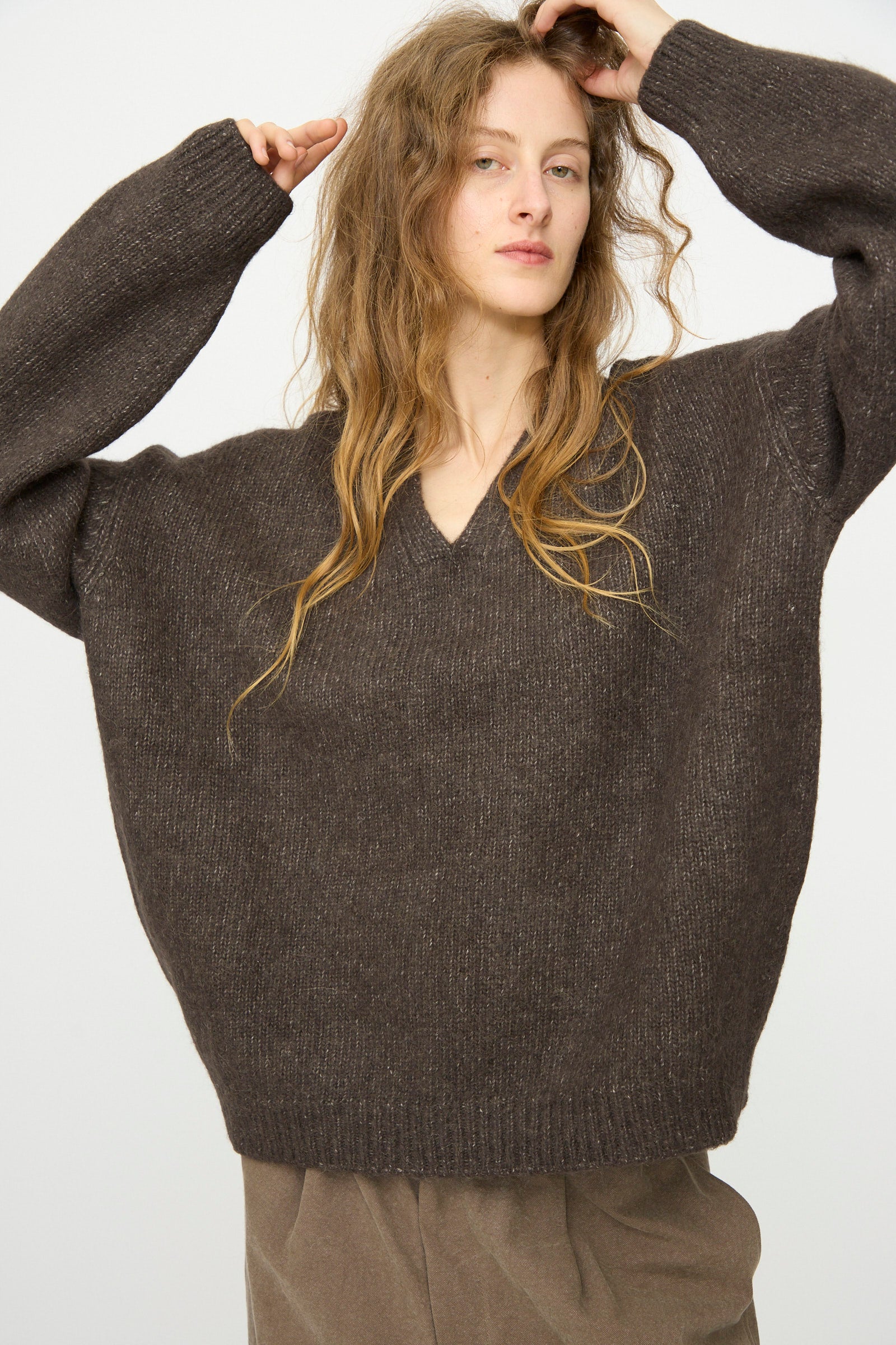 Wearing the Lauren Manoogian Loft V Neck in Coal, a person with long, wavy hair and brown pants stands against a plain light background with arms raised.