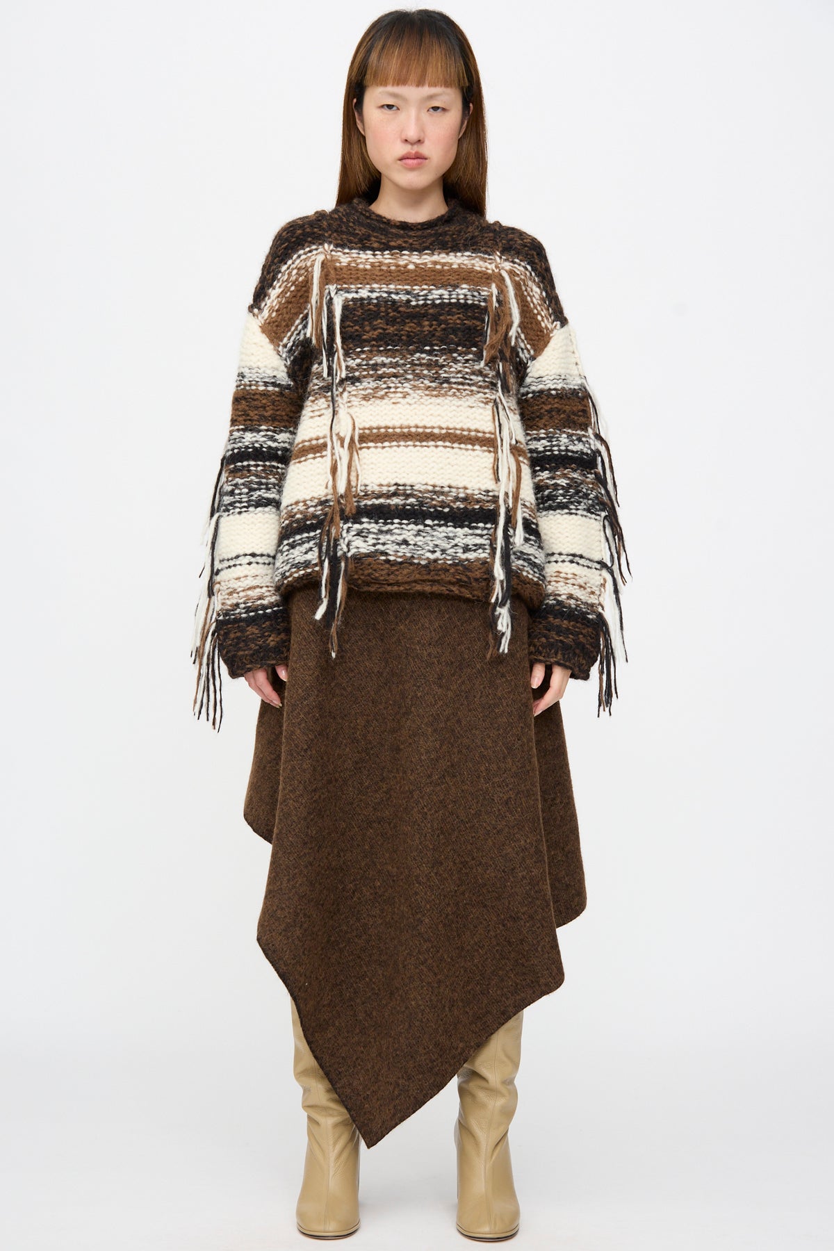 A person wearing a textured brown Lauren Manoogian Manta Skirt in Rust Marl with an asymmetric hem, paired with a striped, fringed knit sweater and tan boots.