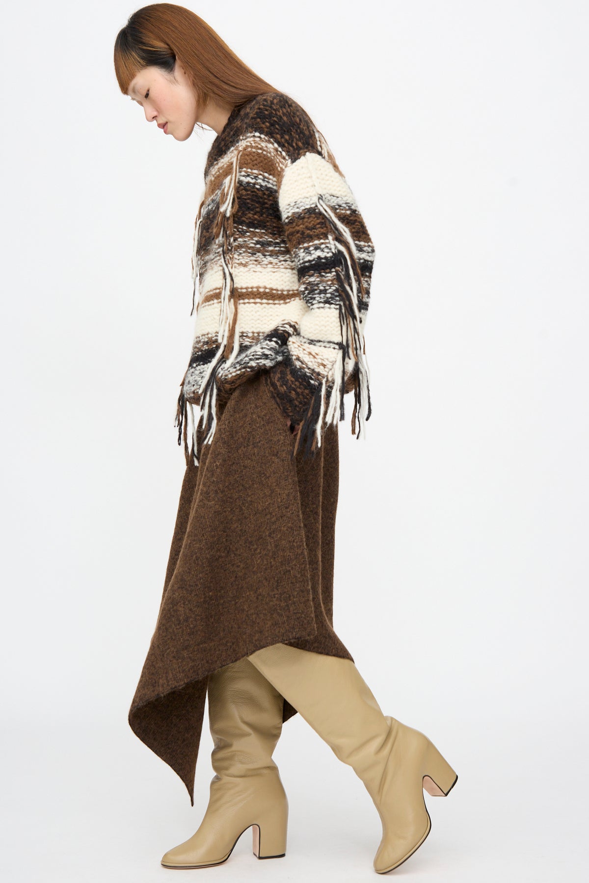 A person wearing a fringed, striped knit sweater and a rust marl Lauren Manoogian Manta Skirt with beige heeled boots.
