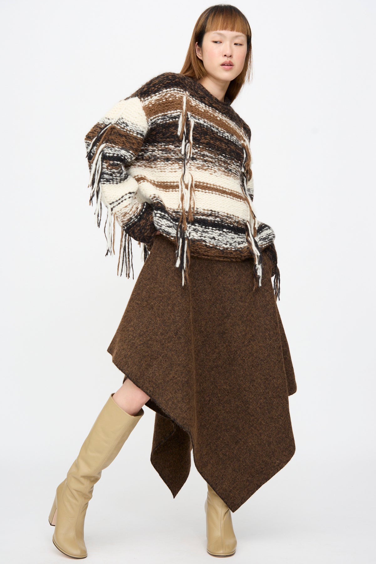 A model wears a brown merino wool Manta Skirt in Rust Marl with a asymmetrical hemline, paired with an oversized striped fringe sweater and beige knee-high boots.