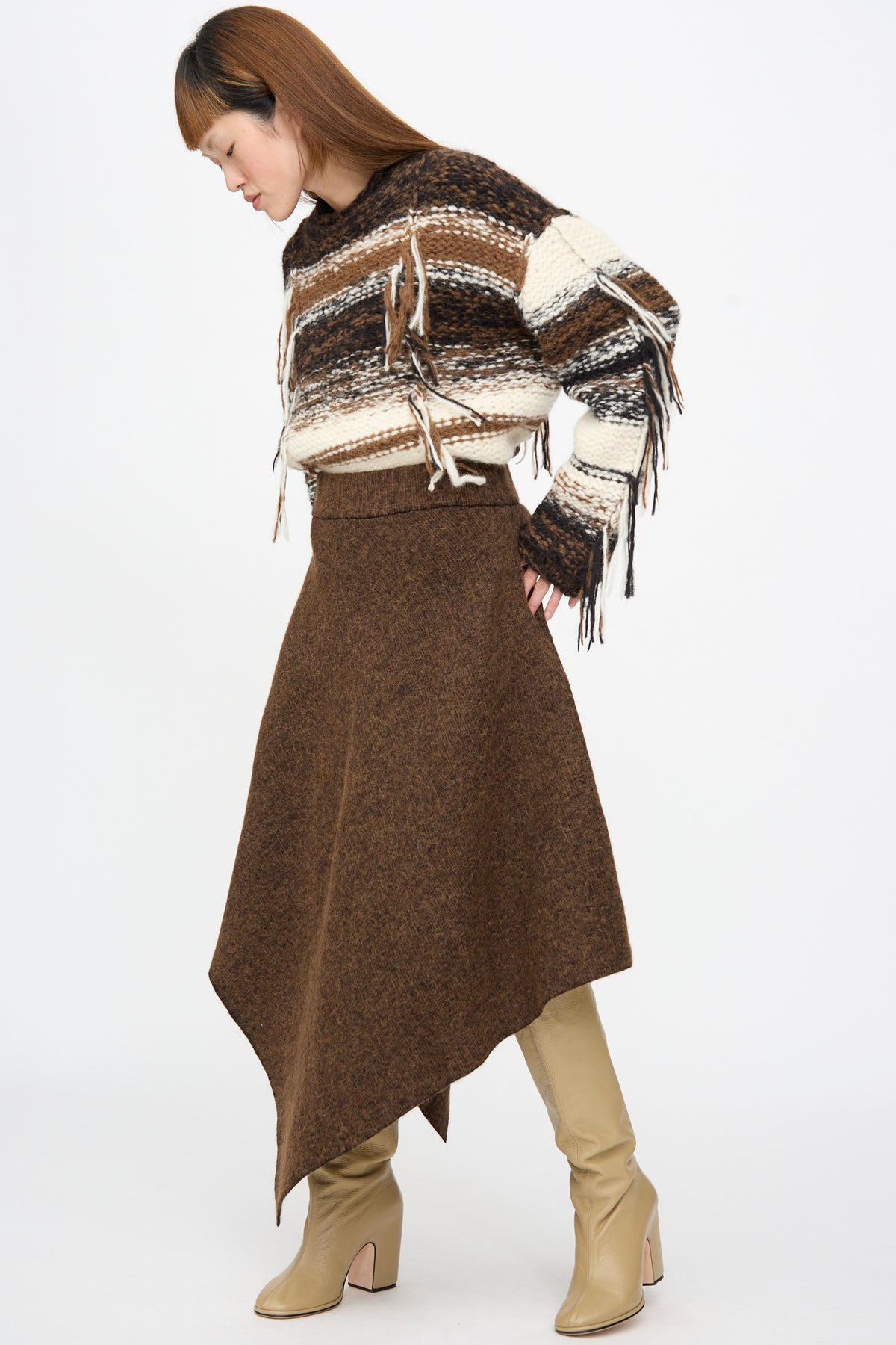 A person wearing a Lauren Manoogian Manta Skirt in Rust Marl, a brown merino wool midi skirt with an asymmetrical hemline, a fringed striped sweater, and tan knee-high boots.