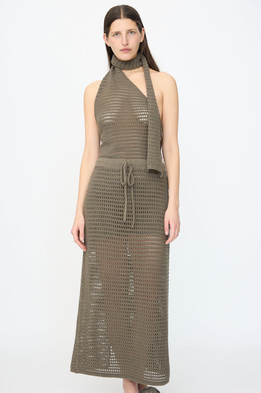 A woman stands against a plain background wearing the Lauren Manoogian Net Skirt in Rock, a sleeveless, olive-green mesh-knit maxi dress crafted from a cotton linen blend, styled with a matching scarf and waist tie.