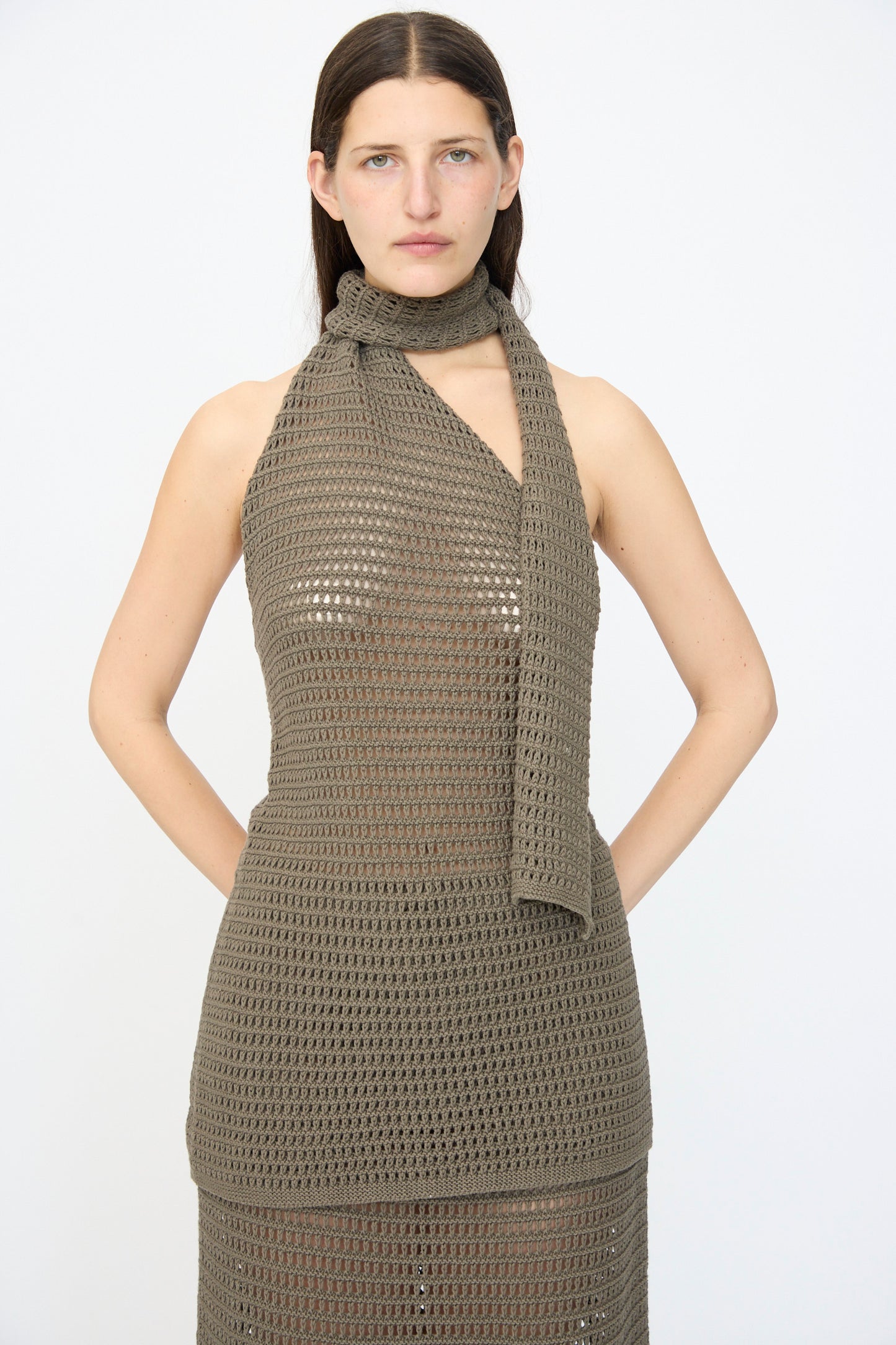 A woman stands against a plain background wearing the Lauren Manoogian Net Wrap Halter in Rock, a sleeveless crochet dress crafted from a cotton linen blend, with a matching crochet scarf draped around her neck.