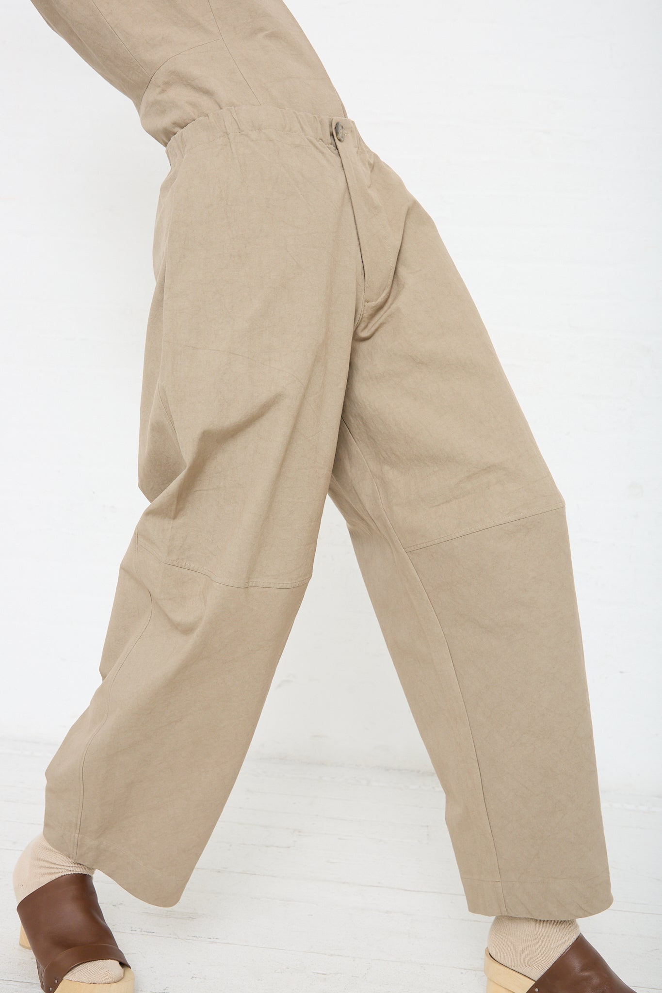 A woman in a New Structure Pant in Drab (Olive brown) by Lauren Manoogian. The model is wearing a matching top and brown sandals. Profile view. 