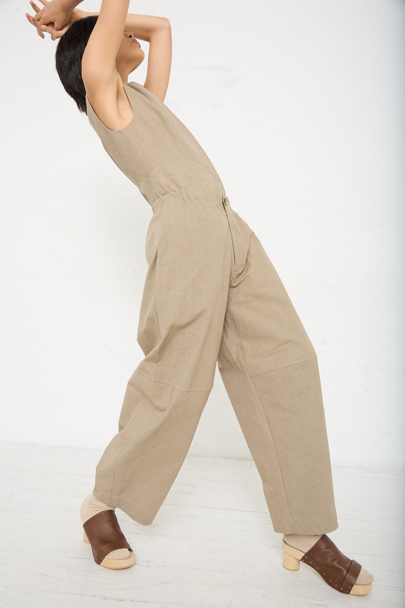 A woman is posing in a New Structure Pant in Drab (Olive Brown) by Lauren Manoogian, which is a relaxed fit cotton pant with an elasticated waist. Full length profile view.