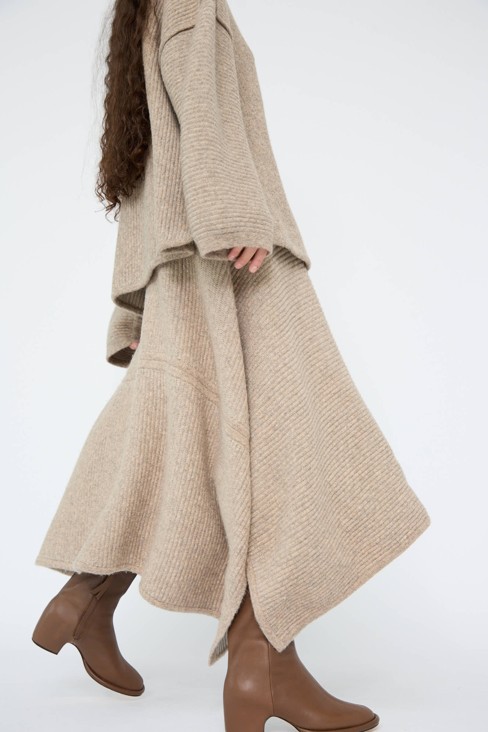 A person wearing a long beige sweater with a draped silhouette, accompanied by the Ottoman Panel Skirt in Moth / Carrara from Lauren Manoogian, effortlessly paired with brown ankle boots.