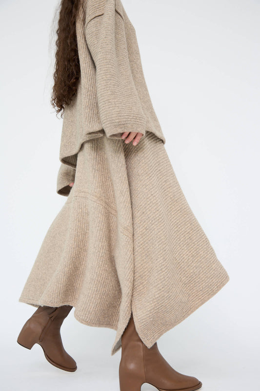 A person wearing a long, beige knit outfit from Lauren Manoogian, specifically the Ottoman Panel Skirt in Moth Carrara with a draped silhouette, pairs it with brown ankle boots as they walk, showcasing their side profile against a plain background.