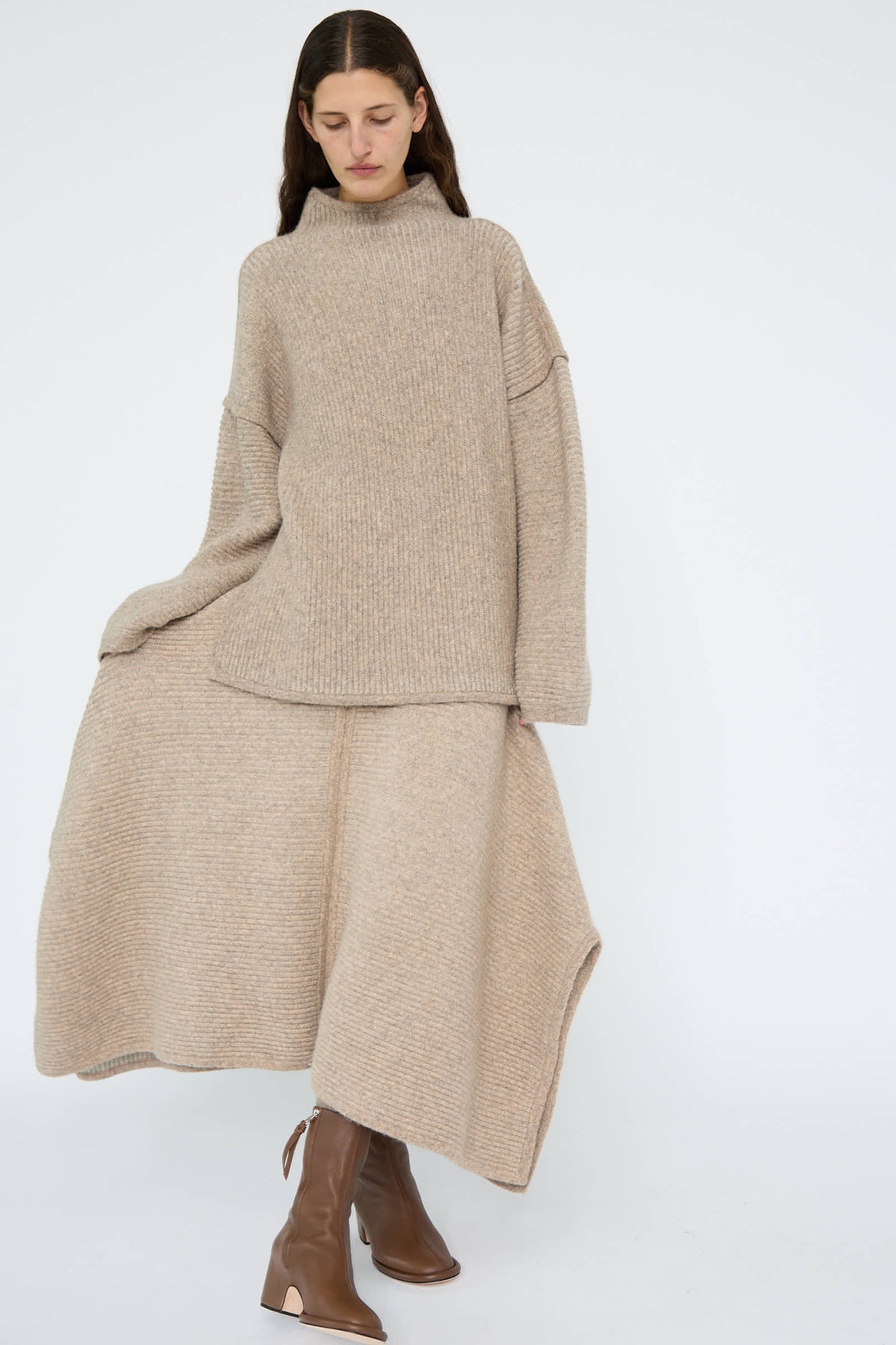 A person wearing a beige knitted sweater and Lauren Manoogian's Ottoman Panel Skirt in Moth / Carrara, crafted from a luxurious wool/alpaca blend, pairs the look effortlessly with brown boots while standing against a plain background.