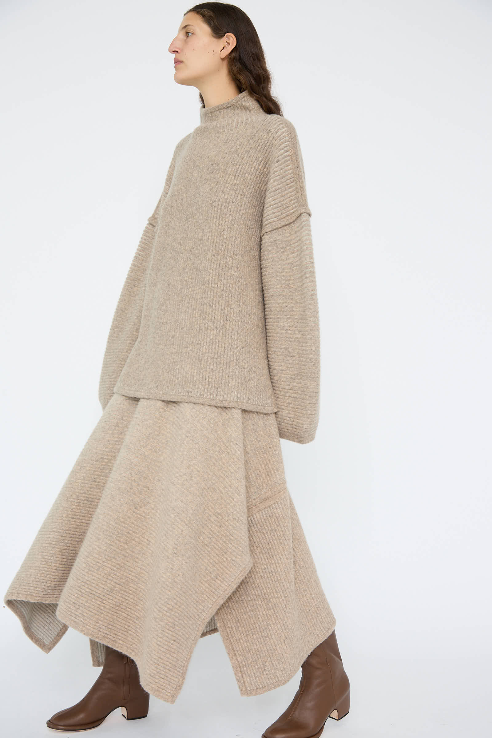 A person stands against a white background, wearing a beige oversized sweater with a draped silhouette and an Ottoman Panel Skirt in Moth / Carrara by Lauren Manoogian, elegantly paired with brown boots.