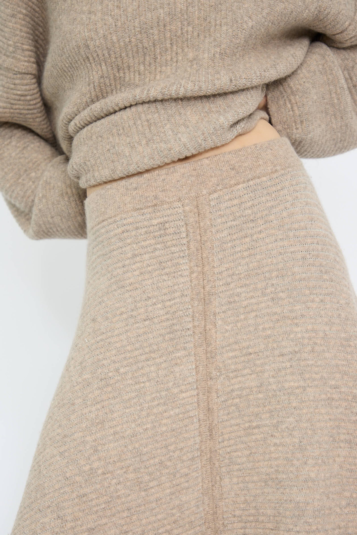 Close-up of a person wearing a beige draped silhouette knit sweater paired with the Ottoman Panel Skirt in Moth/Carrara, designed by Lauren Manoogian, with their hands placed behind their back.