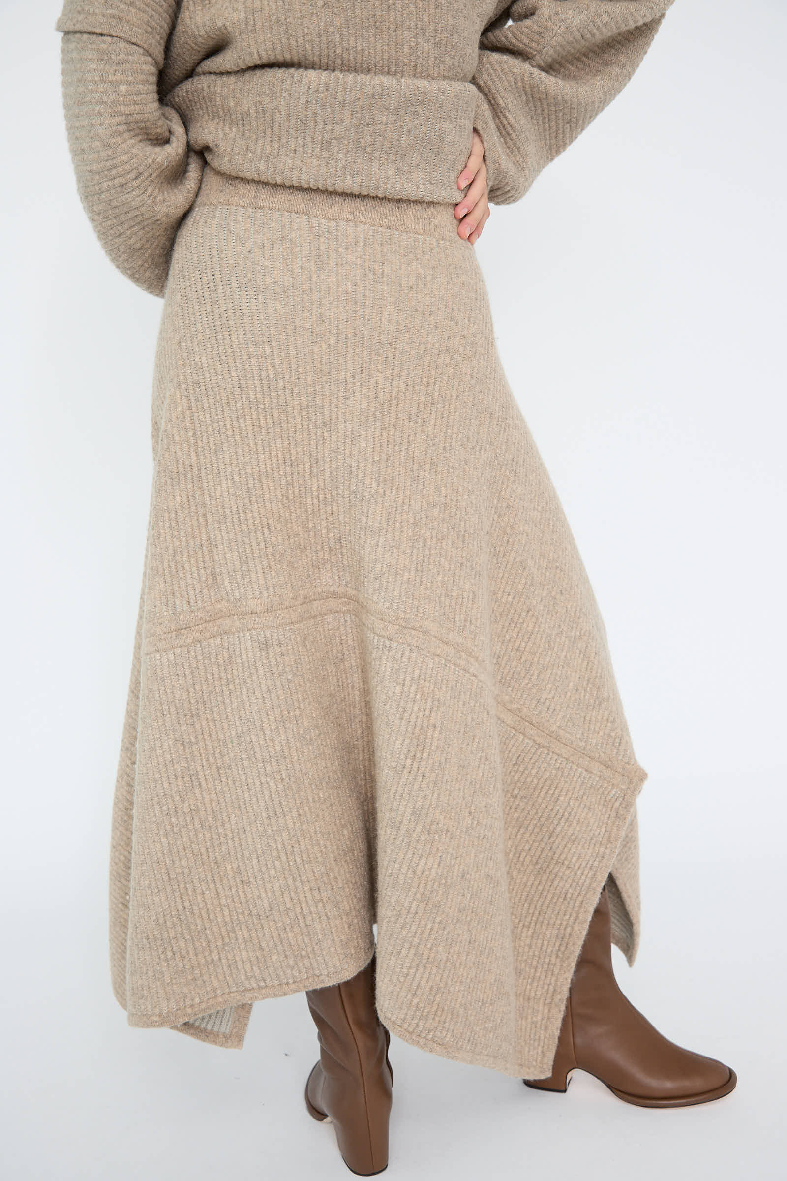 A person is elegantly dressed in the Ottoman Panel Skirt in Moth/Carrara by Lauren Manoogian, with a matching beige knit sweater and brown boots, standing gracefully against a plain background.