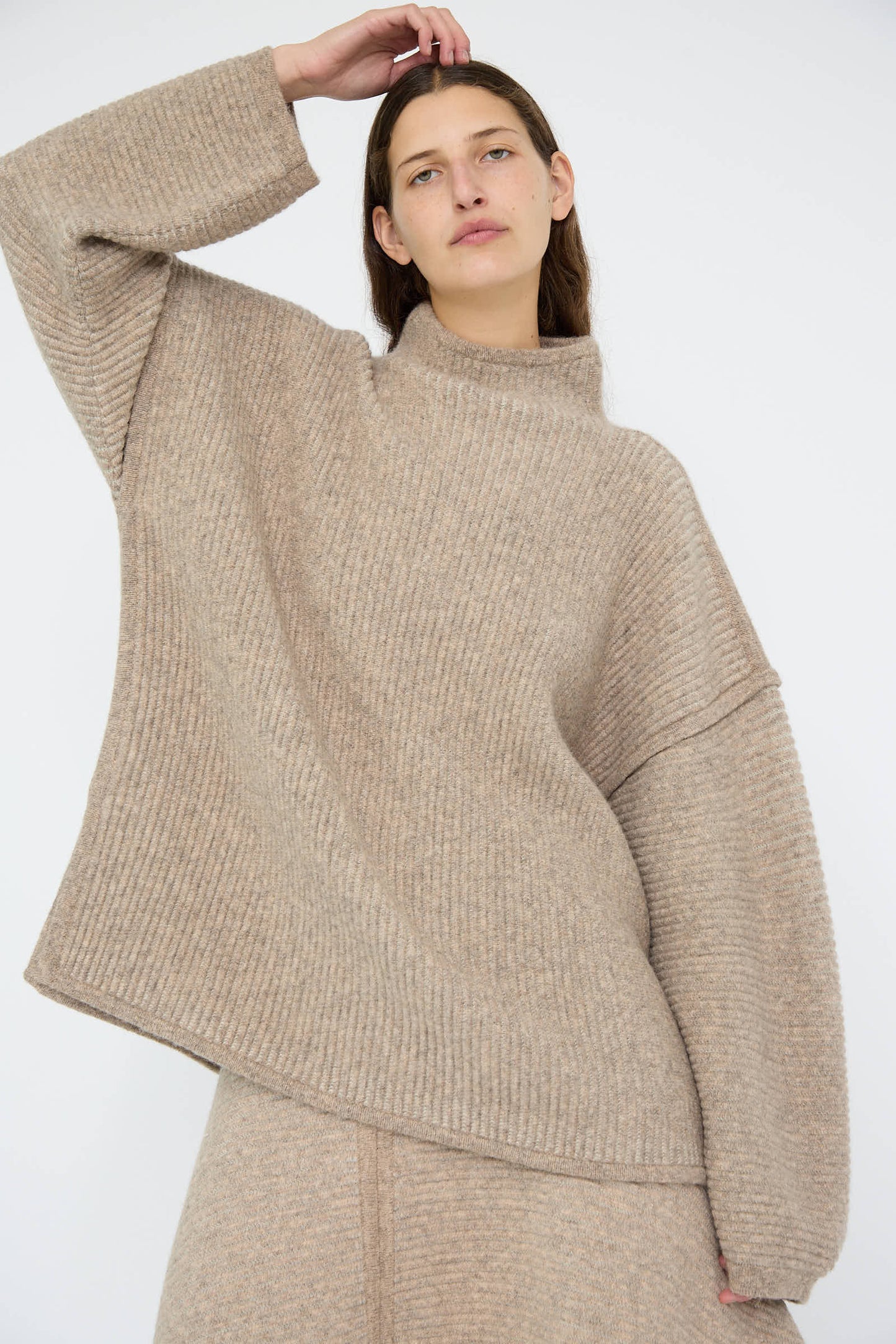 A person is wearing the Ottoman Turtleneck in Moth / Carrara by Lauren Manoogian, a relaxed fit, oversized beige knit sweater crafted from a cozy wool alpaca blend, paired with matching pants against a plain background.