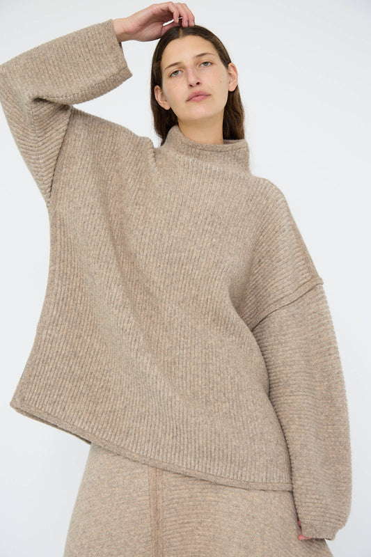 Wearing the Ottoman Turtleneck in Moth Carrara by Lauren Manoogian, the person poses with one arm raised against a plain background, elegantly showcasing the oversized beige wool/alpaca blend sweater and its matching pants.