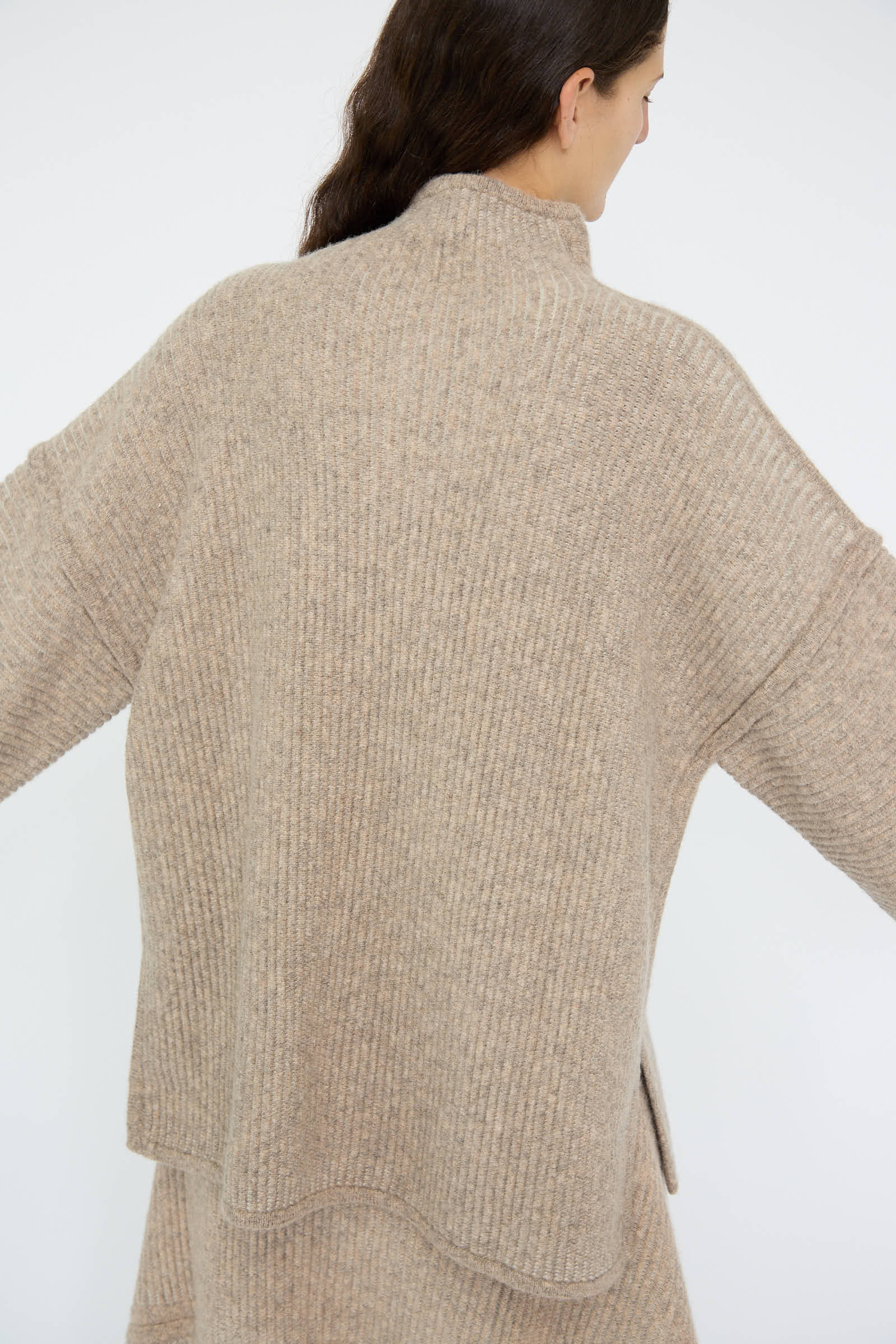 A person wearing the Ottoman Turtleneck in Moth / Carrara, a loose-fitting, beige knitted sweater made from a wool alpaca blend by Lauren Manoogian, is seen from the back with arms slightly extended.