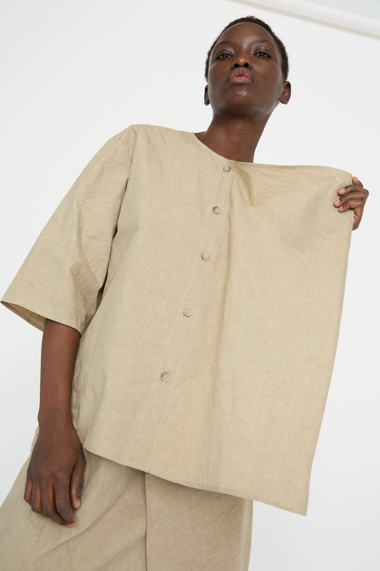 Sporting the Panel Shirt in Drab by Lauren Manoogian, a person in a lightweight, relaxed-fit cotton outfit gently holds the loose-fitting beige shirt to the side as it billows.