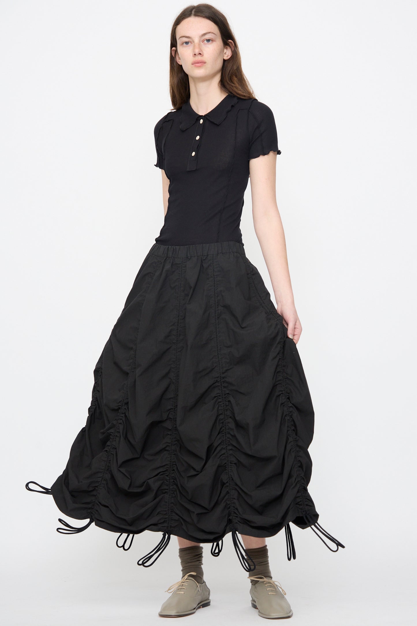 A woman models the Lauren Manoogian Parachute Skirt in Black with a black short-sleeve collared shirt, beige shoes, and gray socks, standing against a plain white background.