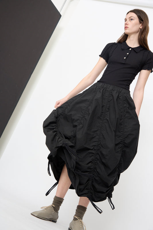 Indoors, a person wears Lauren Manoogian's Parachute Skirt in Black—a cotton midi skirt with an elasticated waistband—paired with a black short-sleeve polo, beige lace-up shoes, and brown socks. 