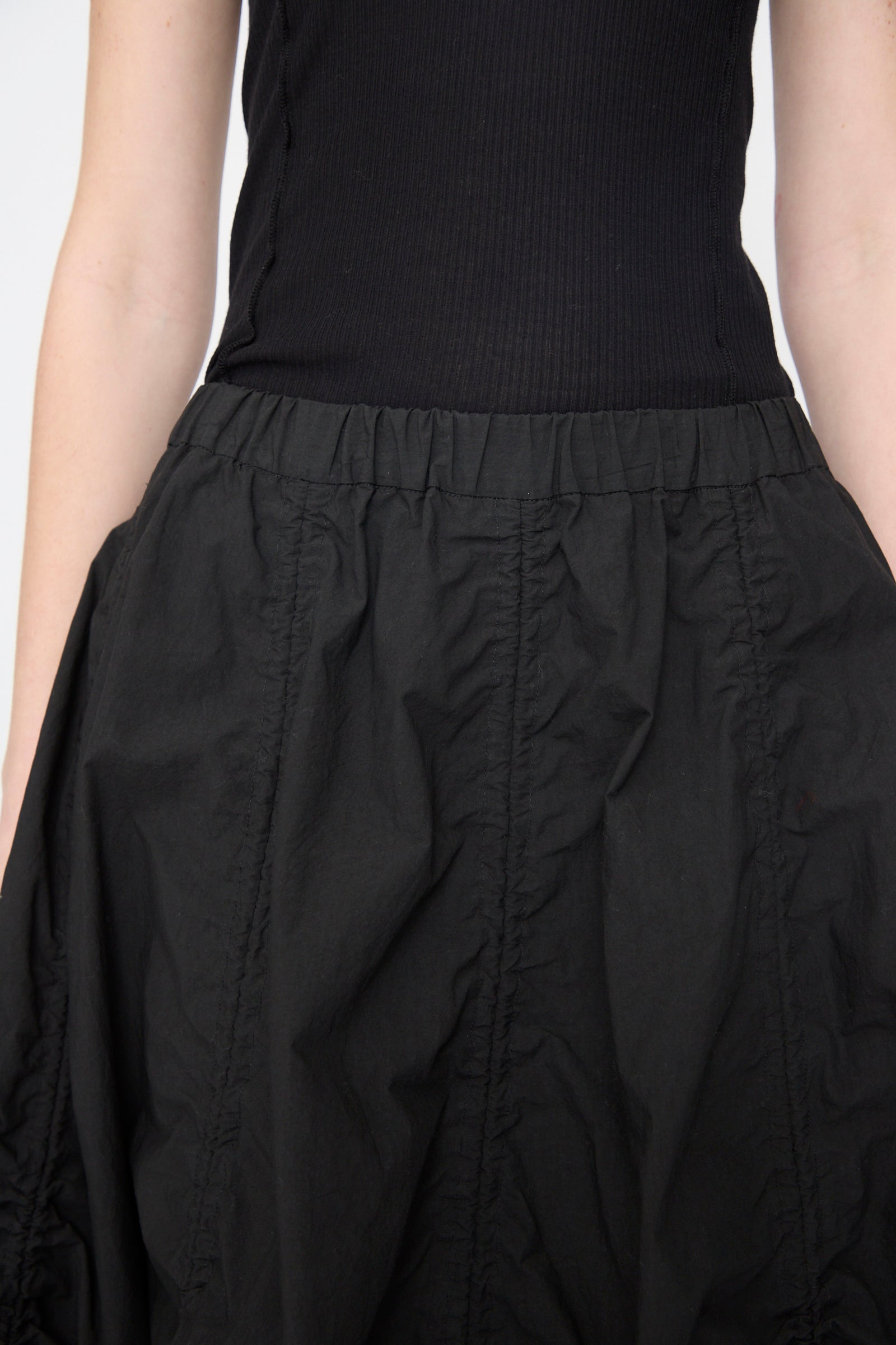 A person wearing a sleeveless black ribbed top and the Lauren Manoogian Parachute Skirt in Black, a high-waisted midi skirt featuring gathered detailing.