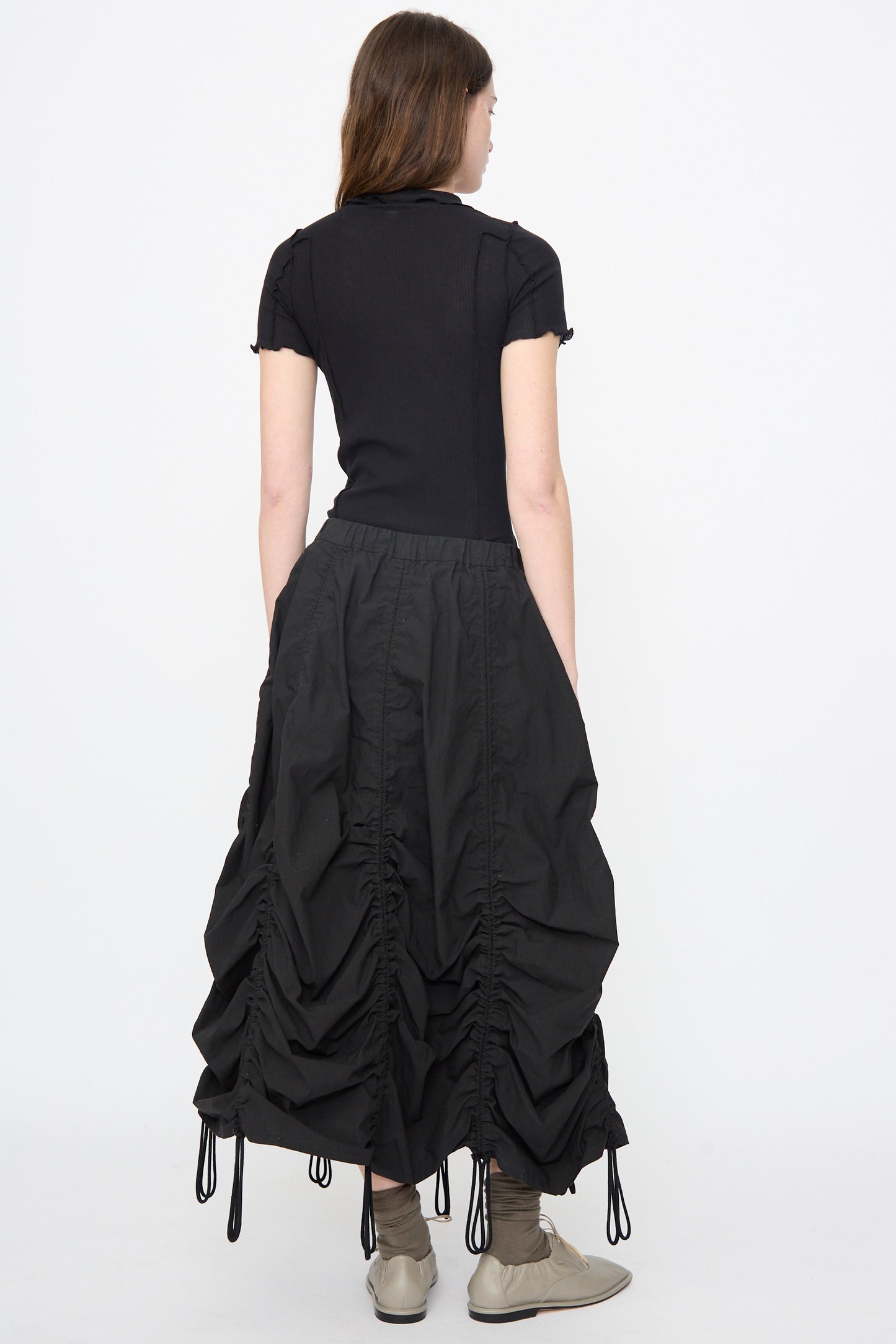 A woman faces away, wearing a short-sleeve black top, Lauren Manoogian’s Parachute Skirt in Black with drawstrings, and beige shoes, set against a plain white background.