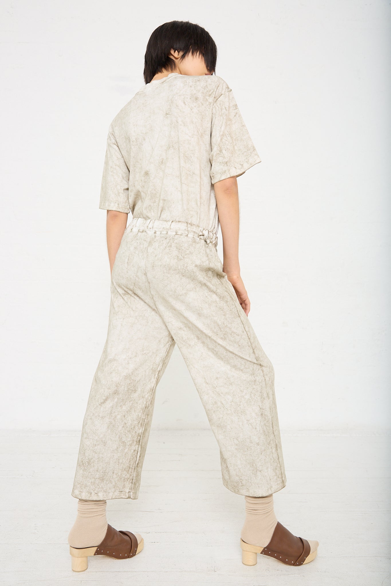A woman is standing in a white room wearing a Pima Cotton Lunar Pant in Olive from Lauren Manoogian. Back view.