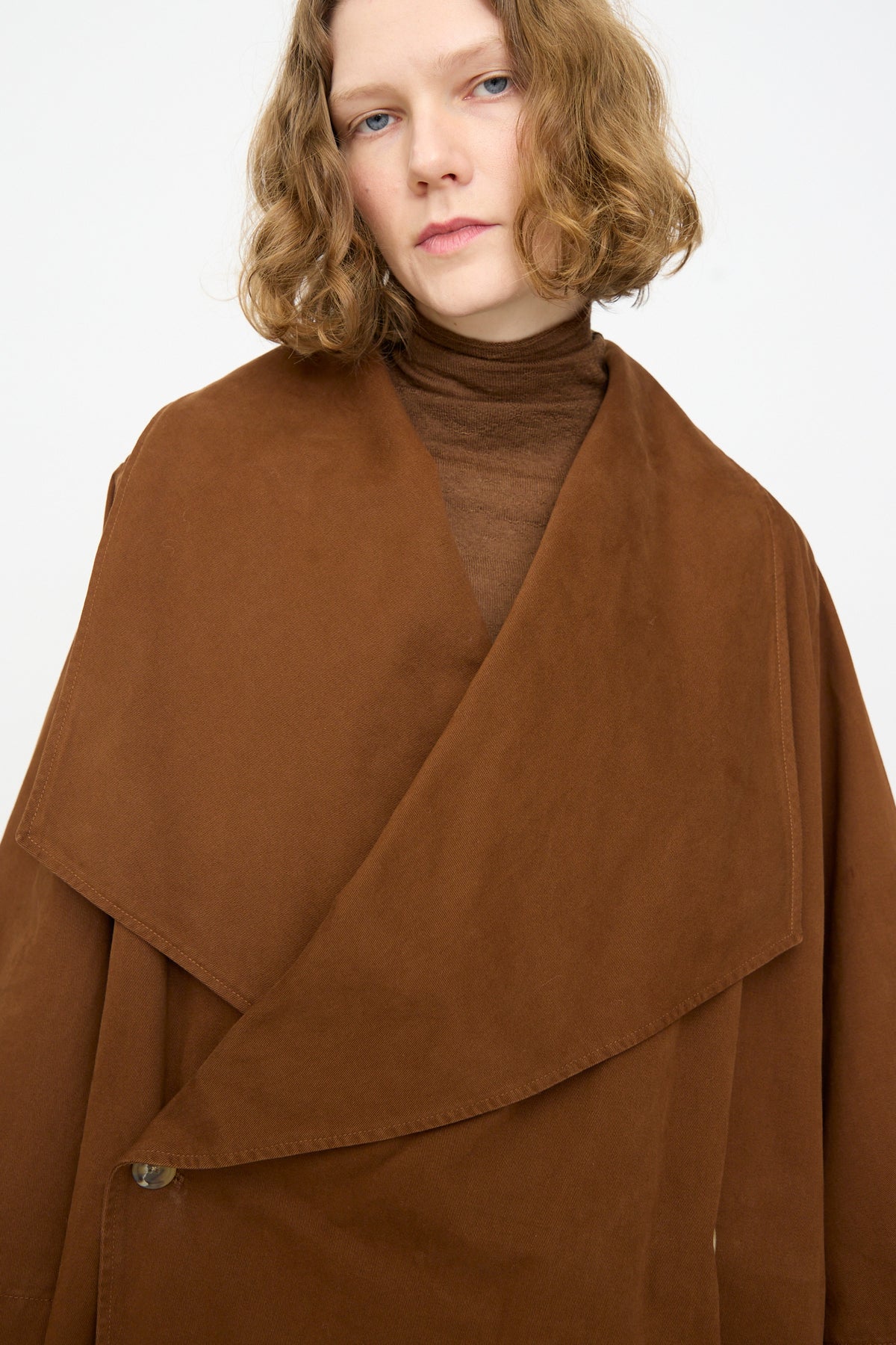 A person with shoulder-length wavy hair wears the Lauren Manoogian Planes Coat in Rust over a brown turtleneck, standing against a plain white background.
