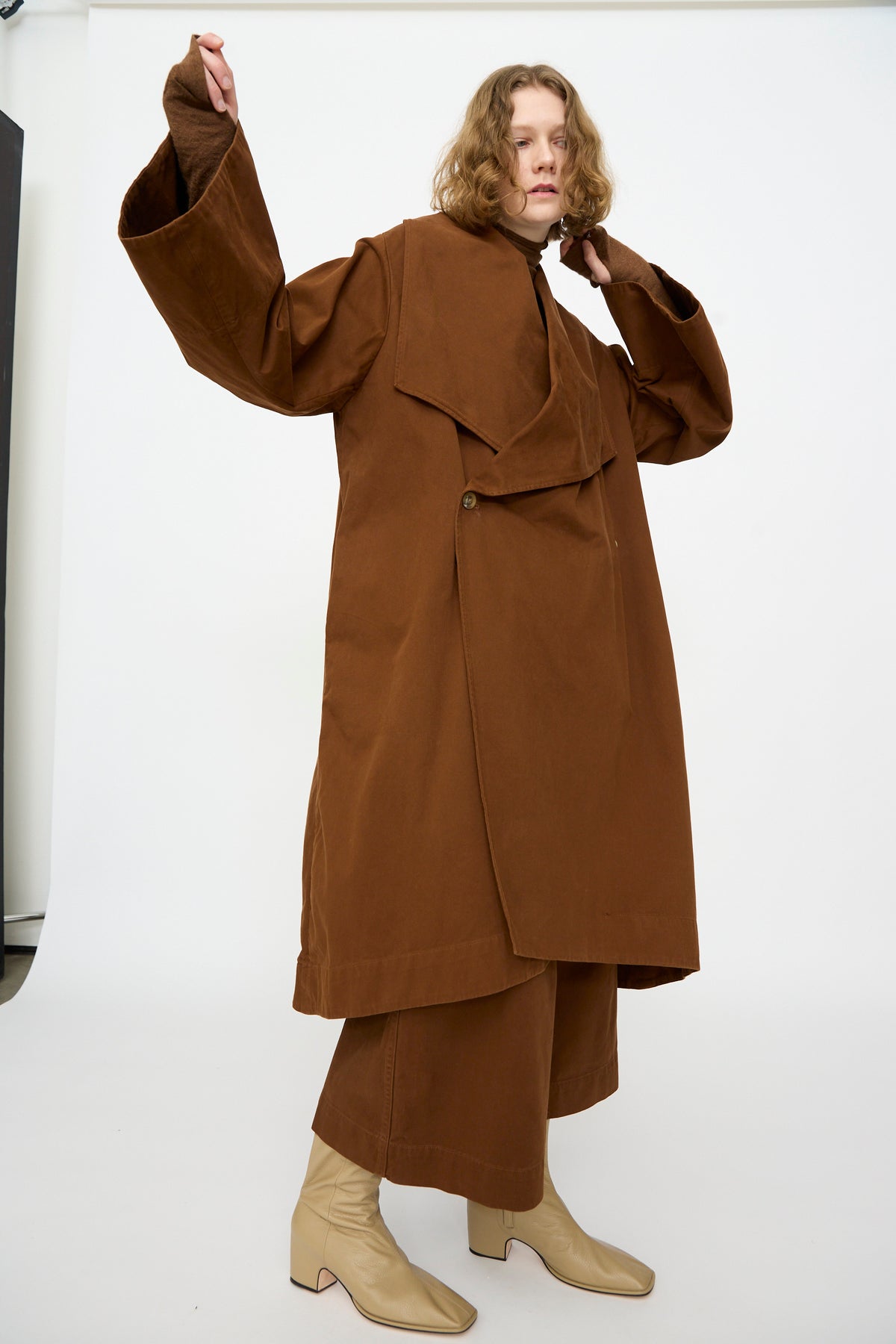 A person wearing the Lauren Manoogian Planes Coat in Rust— a loose-fitting, mid-length cotton coat— and wide brown pants, paired with beige heeled boots, stands against a plain white background.
