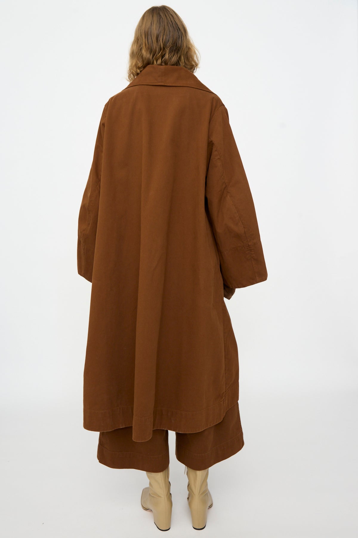 Wearing the Lauren Manoogian Planes Coat in Rust, paired with matching brown pants and cream-colored boots, the person stands with their back to the camera against a plain white background.
