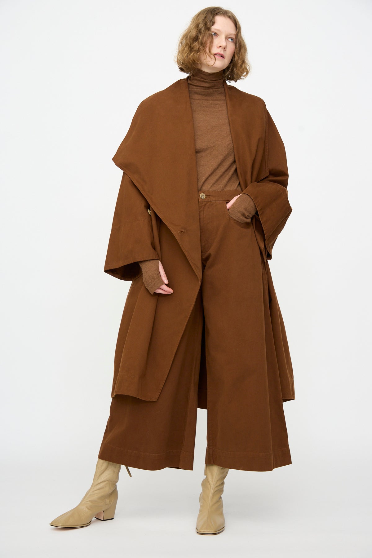 A person wearing the Lauren Manoogian Planes Coat in Rust—an oversized, long cotton coat—paired with wide-leg brown pants, a brown turtleneck, and beige boots, standing against a plain white background.