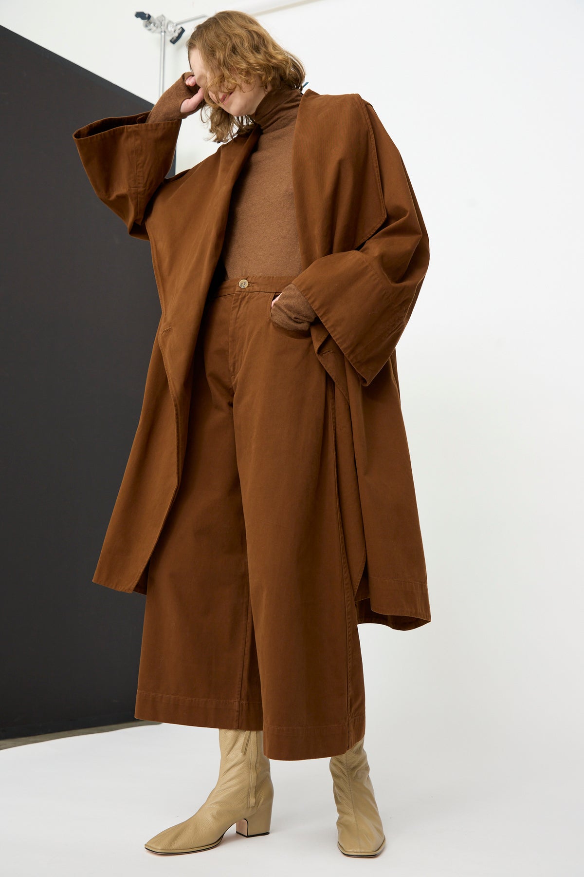 A person wearing the Lauren Manoogian Planes Coat in Rust, styled with a brown turtleneck sweater, wide-leg brown pants, and beige boots, stands against a white and black background.