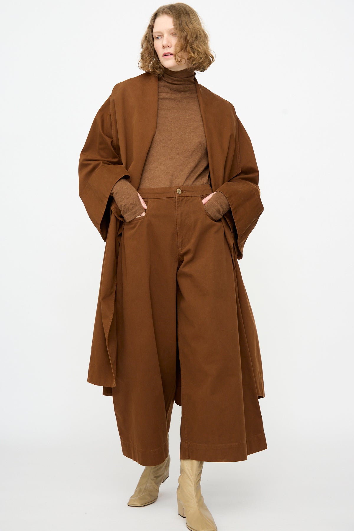 A person wearing the Lauren Manoogian Planes Coat in Rust, styled with a brown turtleneck sweater, wide brown pants, and beige ankle boots, standing against a plain white background.